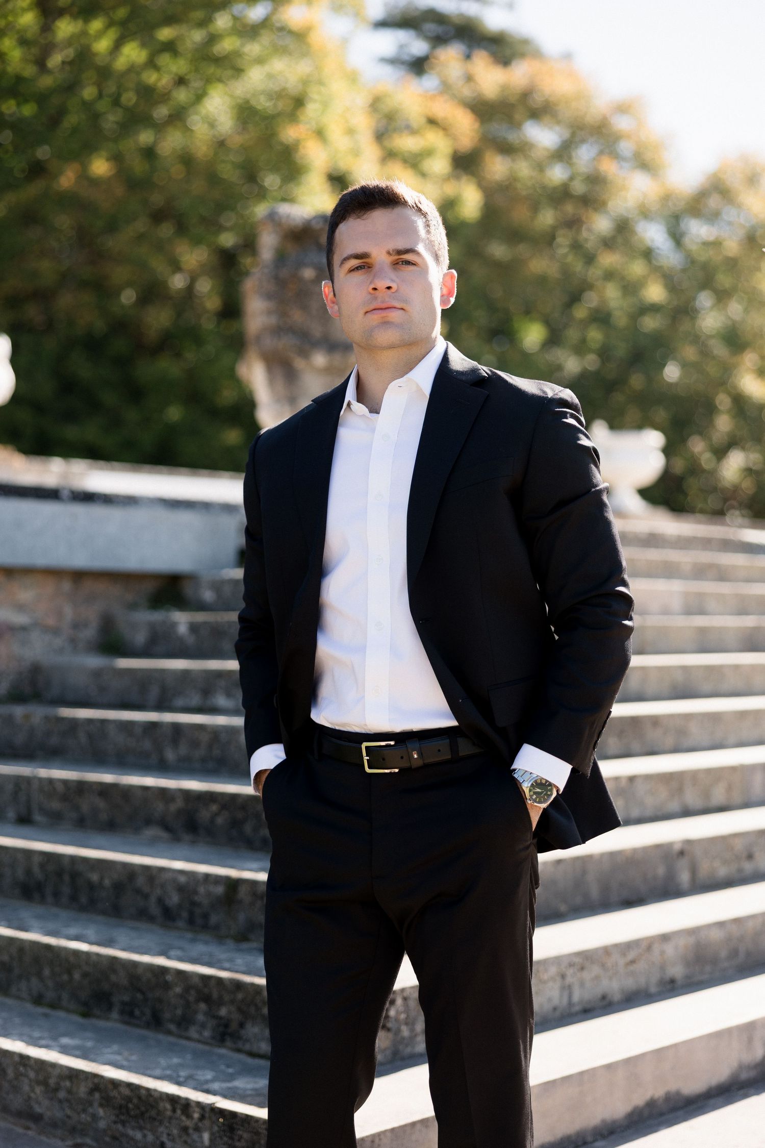 Confident groom in black suit standing on stone steps surrounded by autumn trees in a château garden.