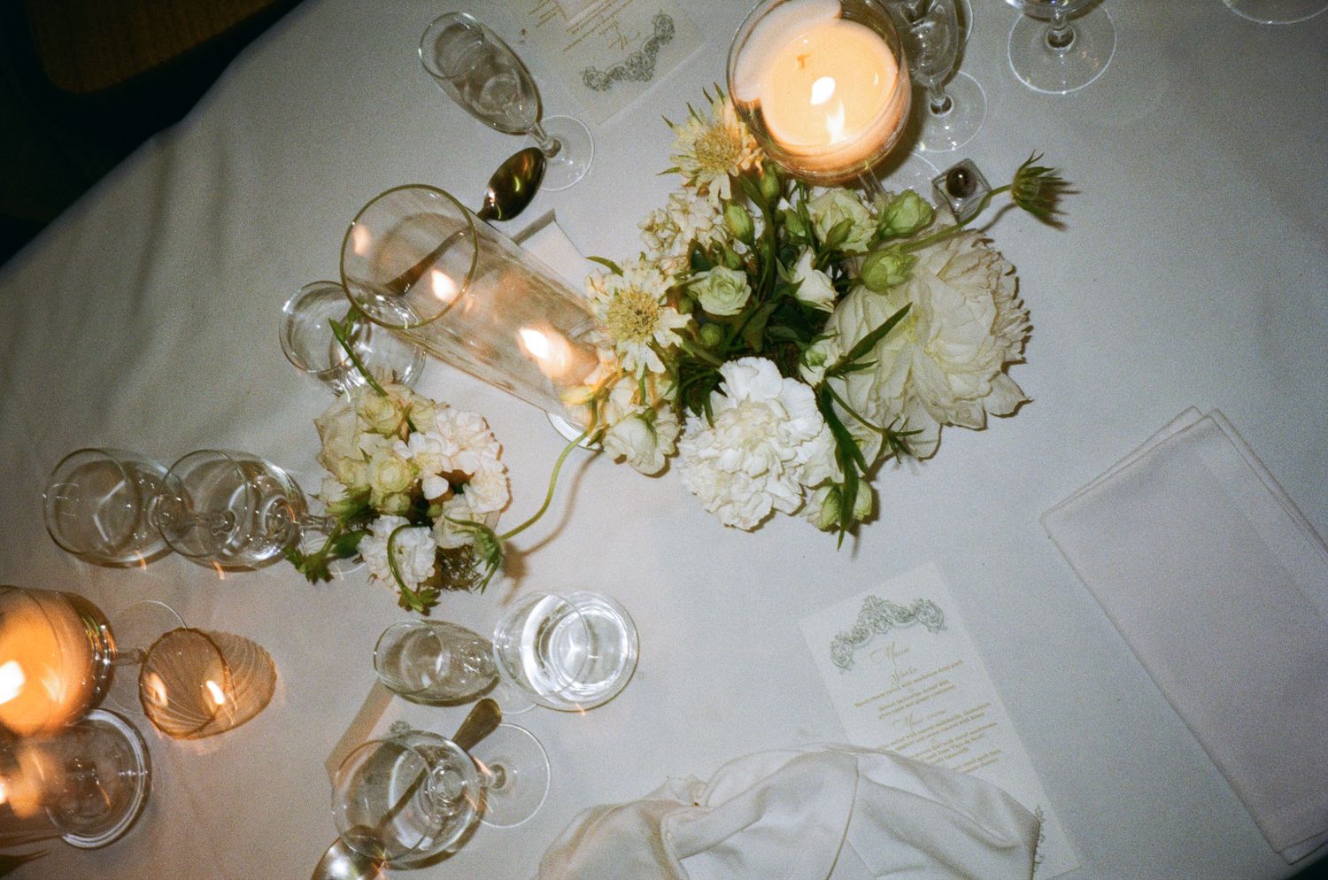 Elegant wedding reception table setting with glowing candles, white flowers and crystal glassware on a white tablecloth.