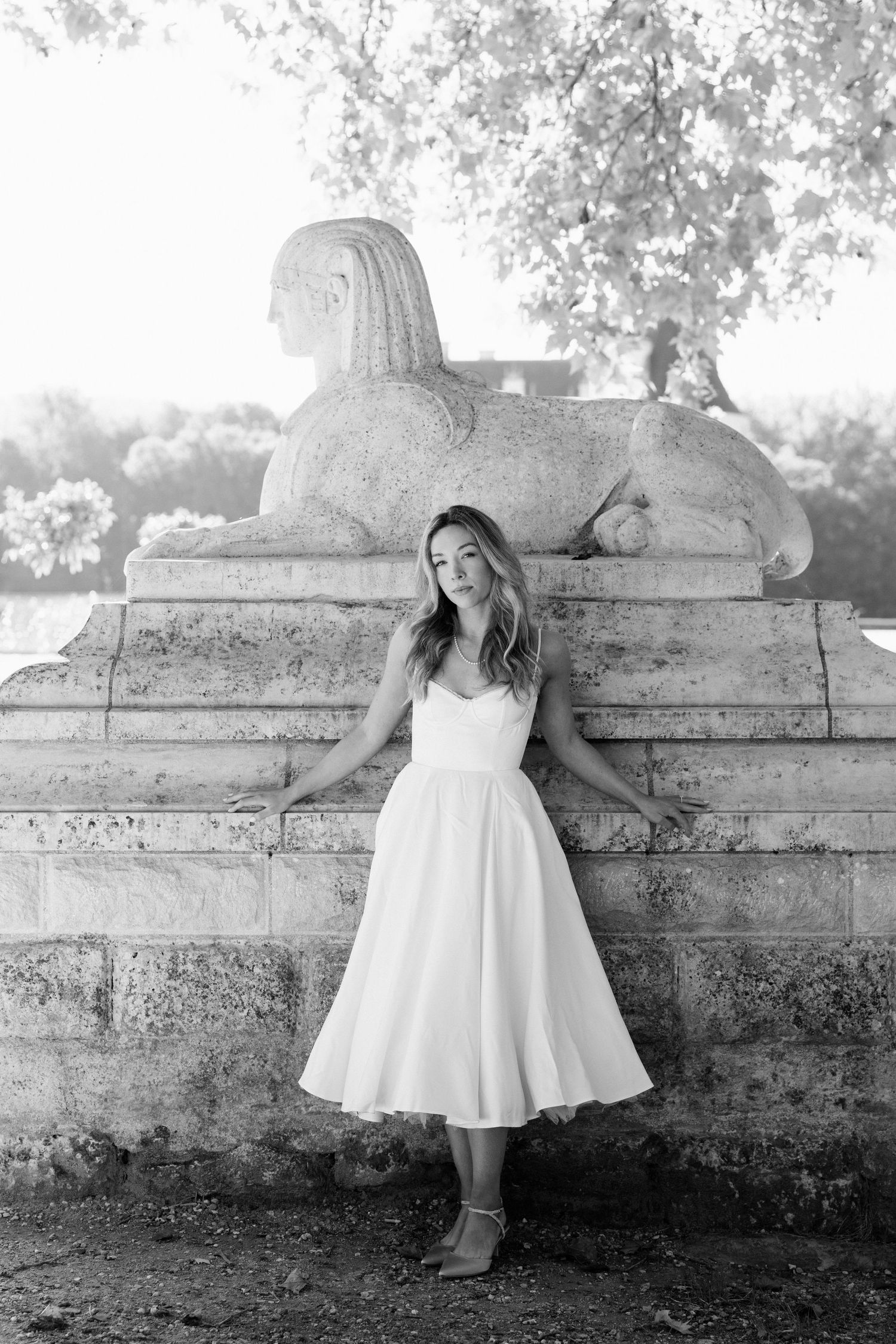 Black and white portrait of a bride in white midi dress leaning against a stone sphinx sculpture outdoors.
