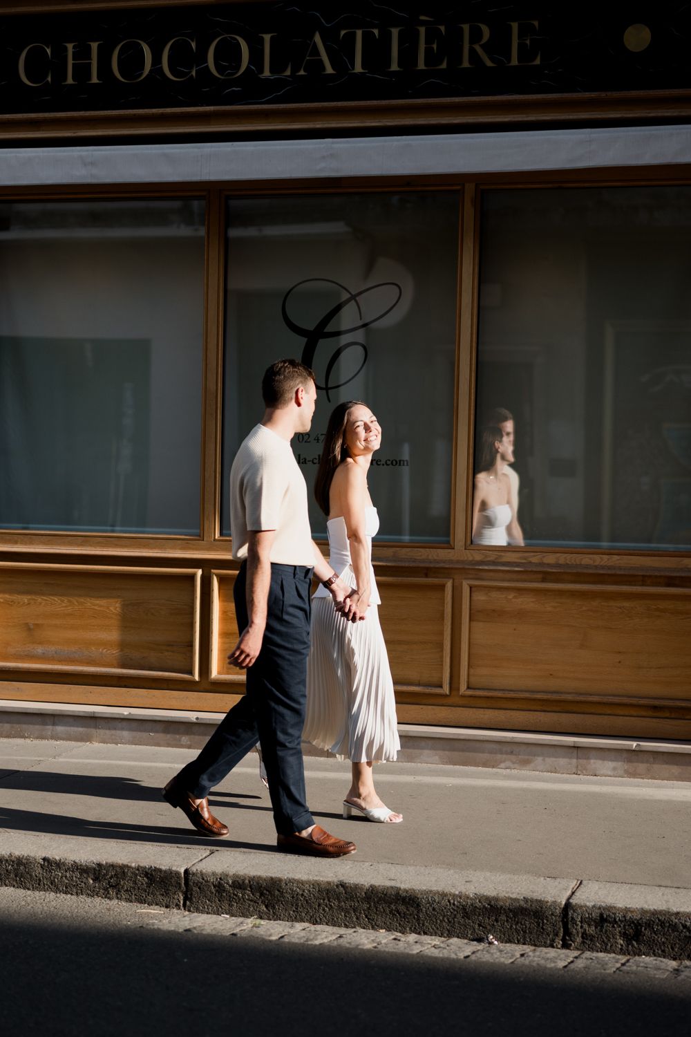 Couple walking past a storefront with 'Chocolatiere' signage in soft natural lighting.