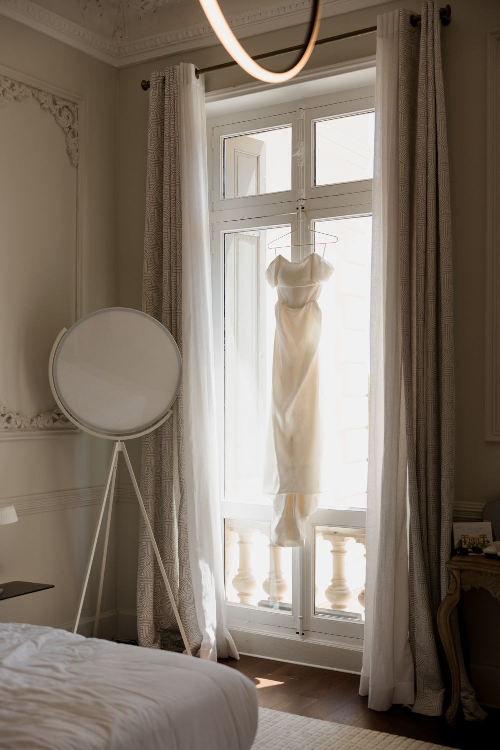 Elegant white wedding dress hanging in sunlit window with flowing curtains and minimalist modern decor in bedroom.