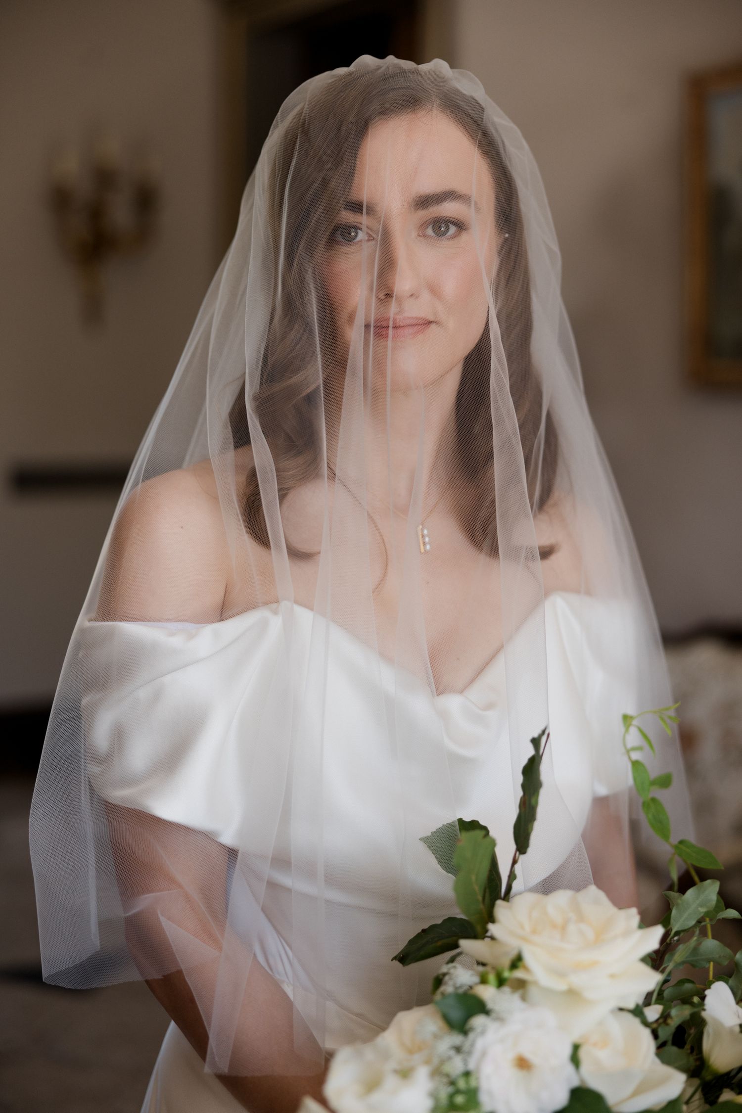 A bride in a white wedding dress holds a bouquet of white roses while wearing a flowing veil in a soft-lit indoor setting.