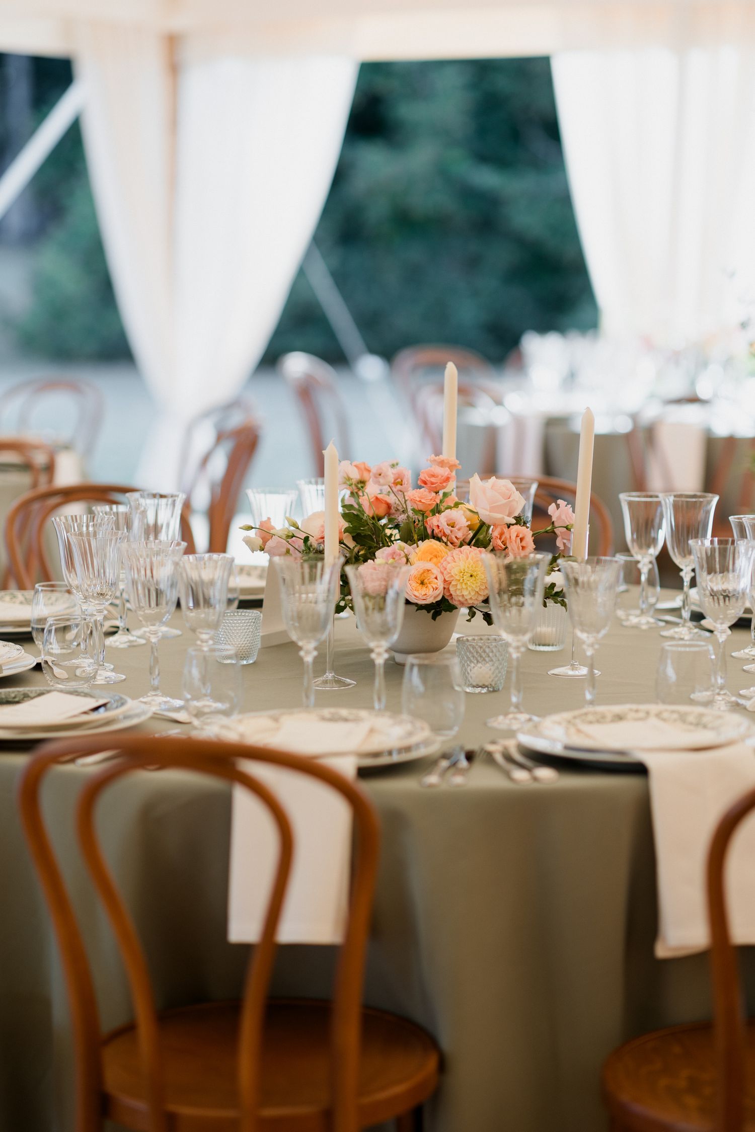 Elegant wedding reception table settings with peach floral centerpieces and wooden chairs under a white tent.