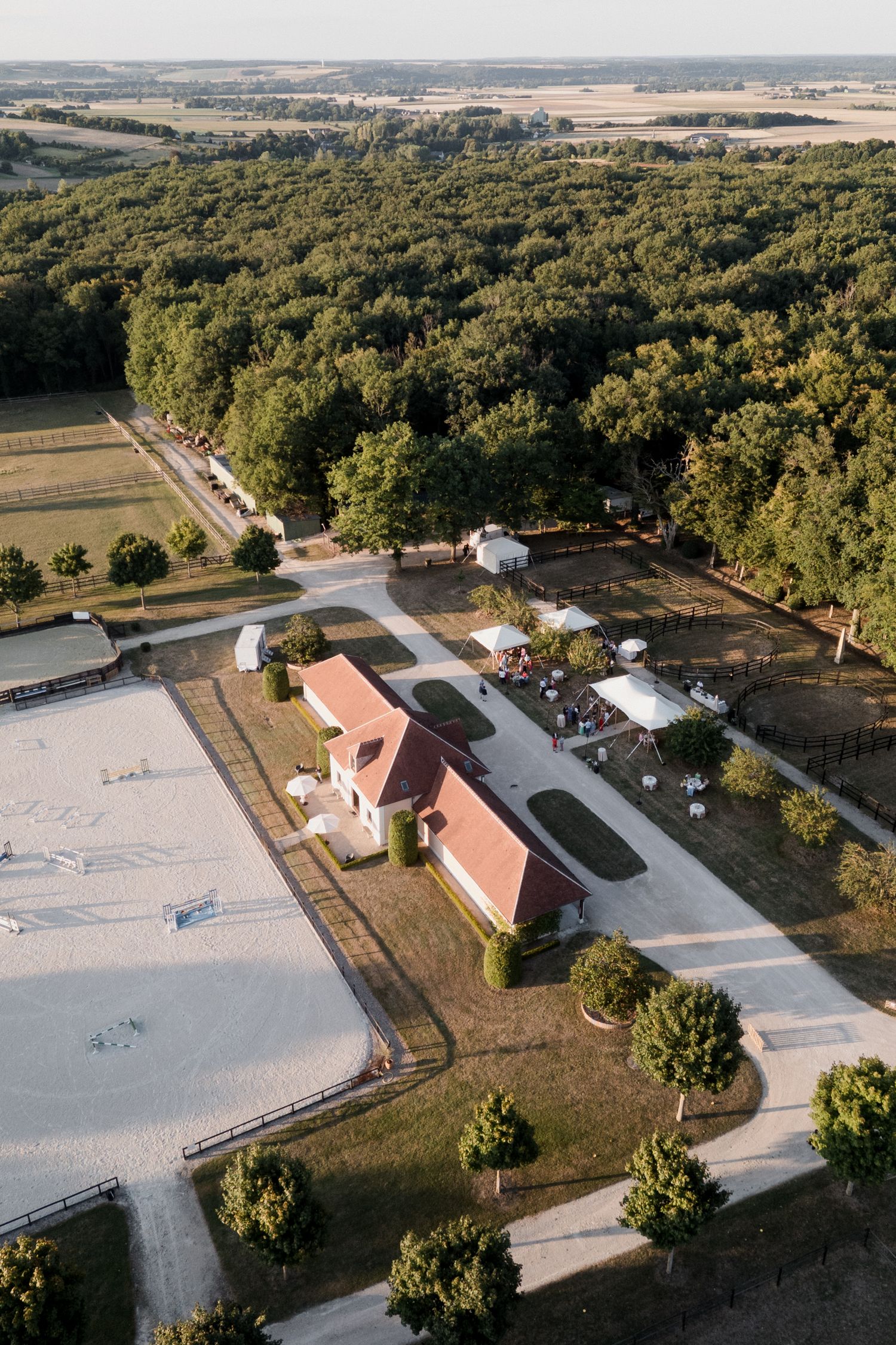 Aerial view of an equestrian facility with riding arena, barn buildings, and surrounding wooded countryside.