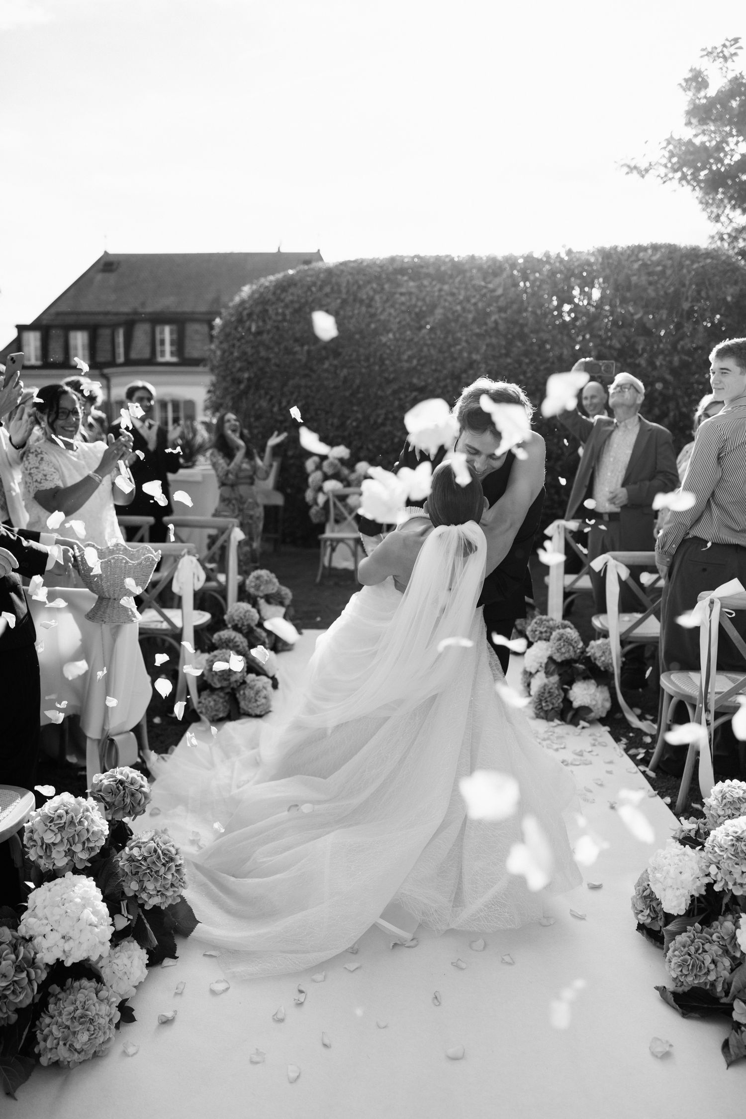 Bride and groom walk down outdoor aisle as guests toss white flower petals during wedding ceremony.