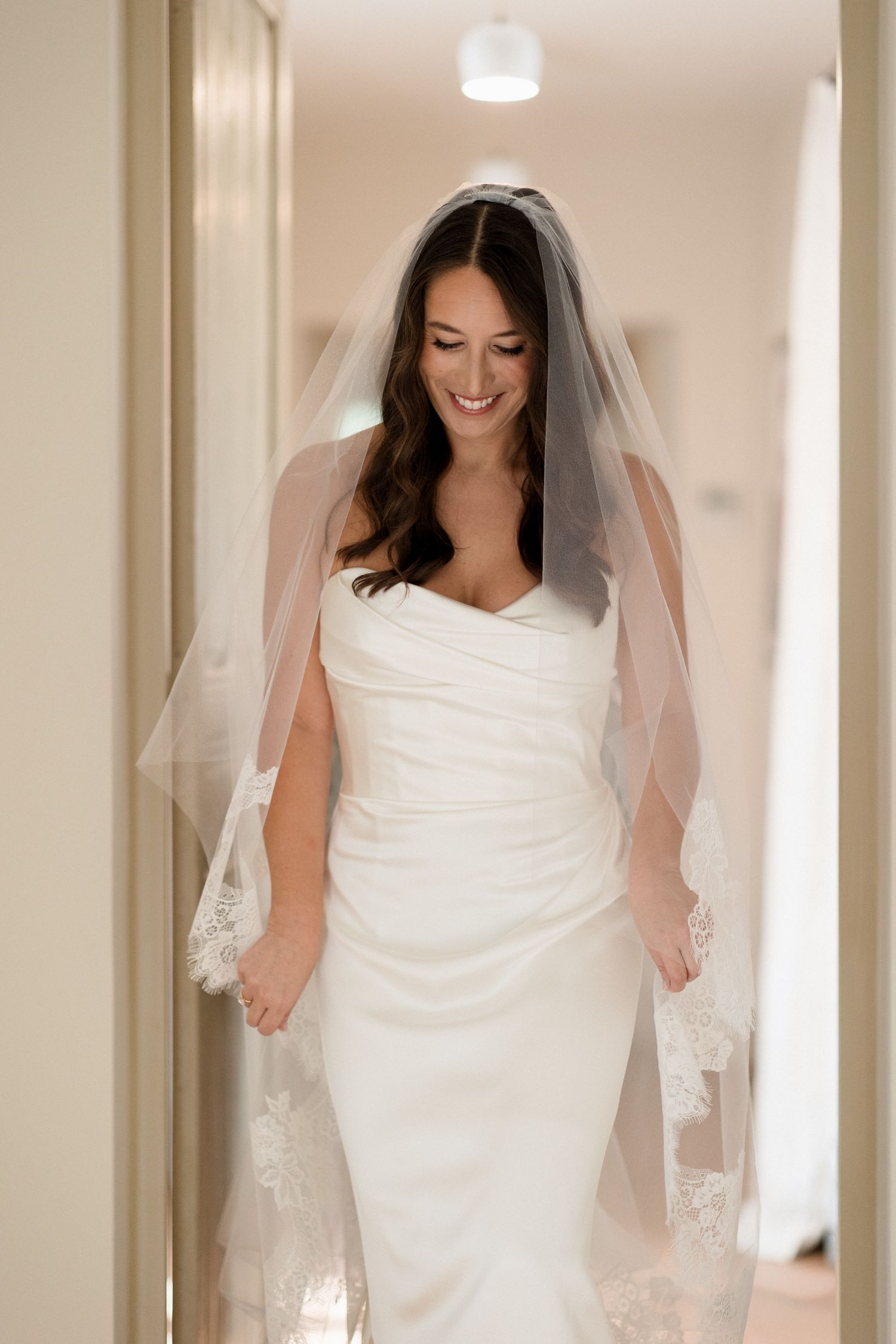 A bride in a white wedding gown and veil walks down a brightly lit hallway.