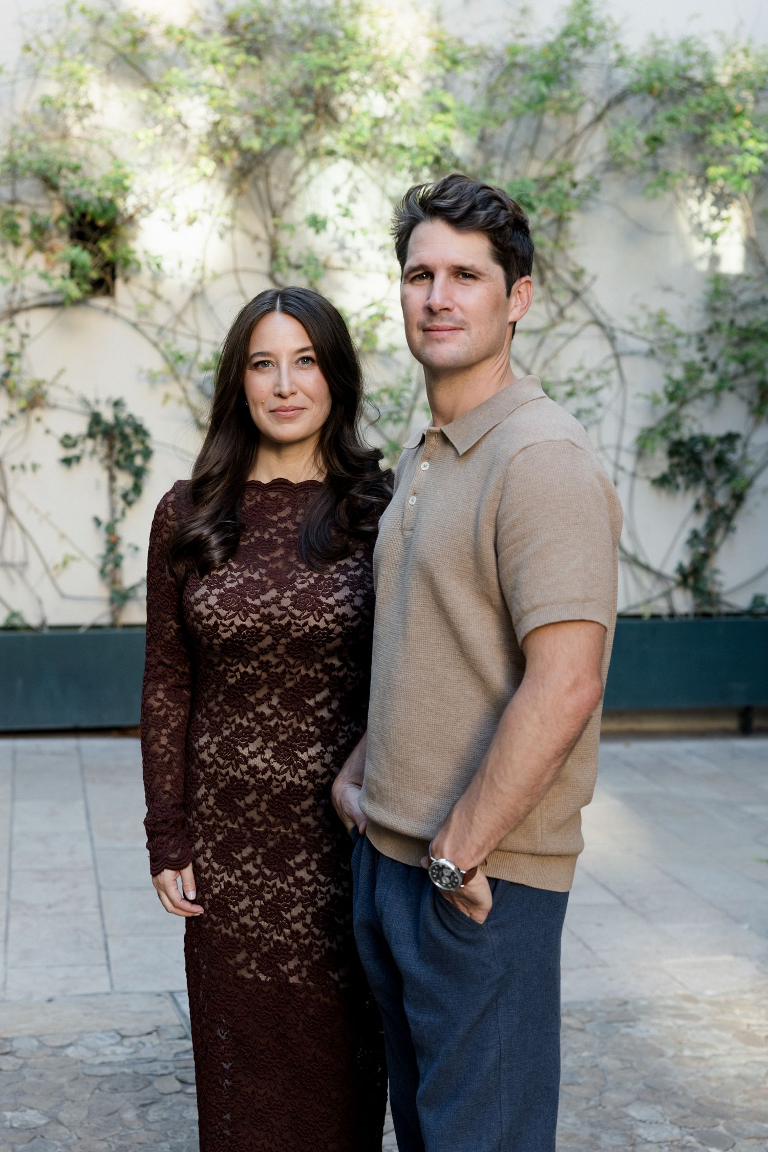 A couple poses together against a white wall with climbing vines, wearing brown and blue casual attire. Engagement session Paris