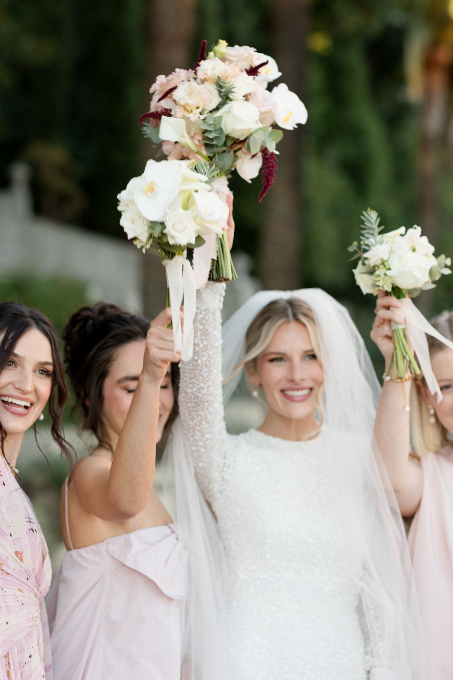 A group of bridesmaids in white and pink attire hold up matching white floral bouquets while celebrating at a wedding.