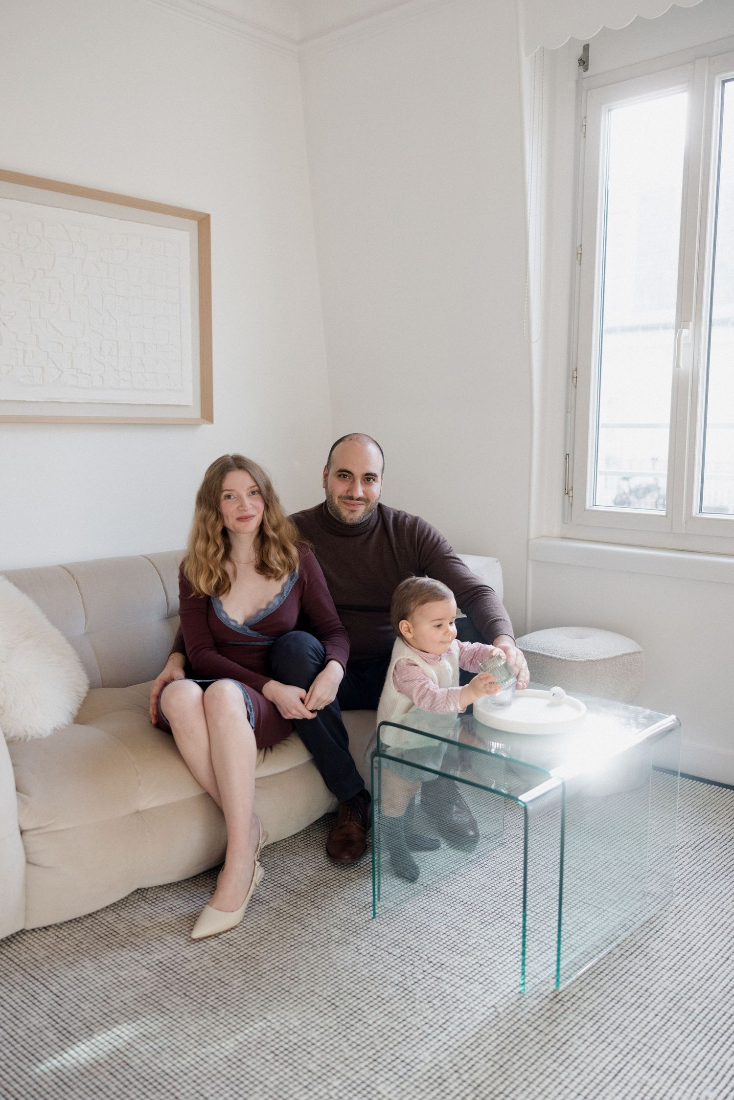 A young family with a toddler sits on a beige sofa in a bright, minimalist living room with a glass coffee table.