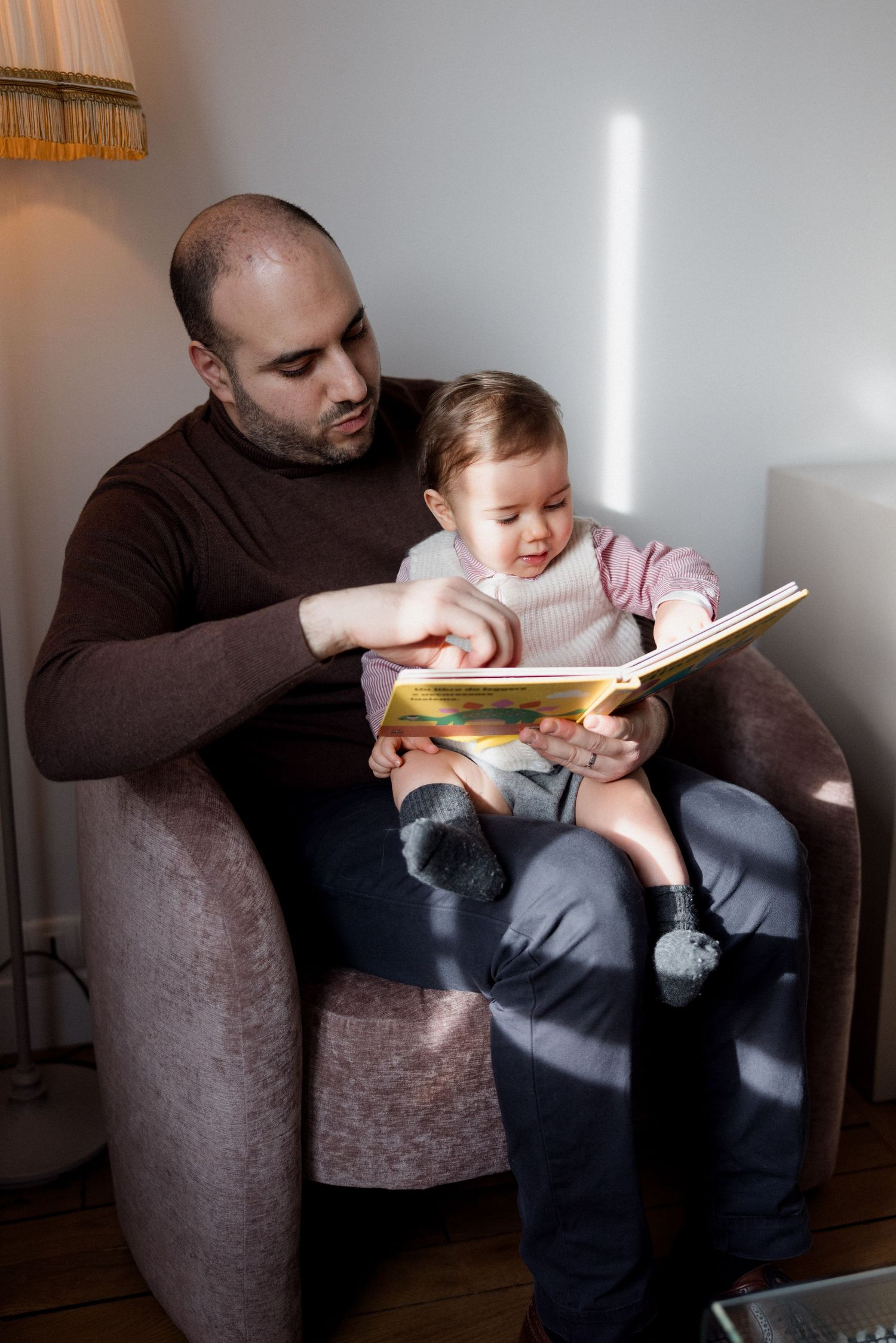 Father sitting in armchair reading a colorful book with his baby daughter on his lap at home.