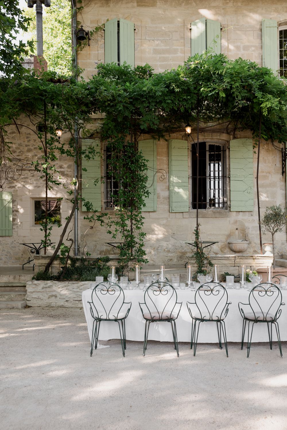 Rustic French countryside villa with sage green shutters and climbing vines features white metal garden chairs against stone walls. Avignon