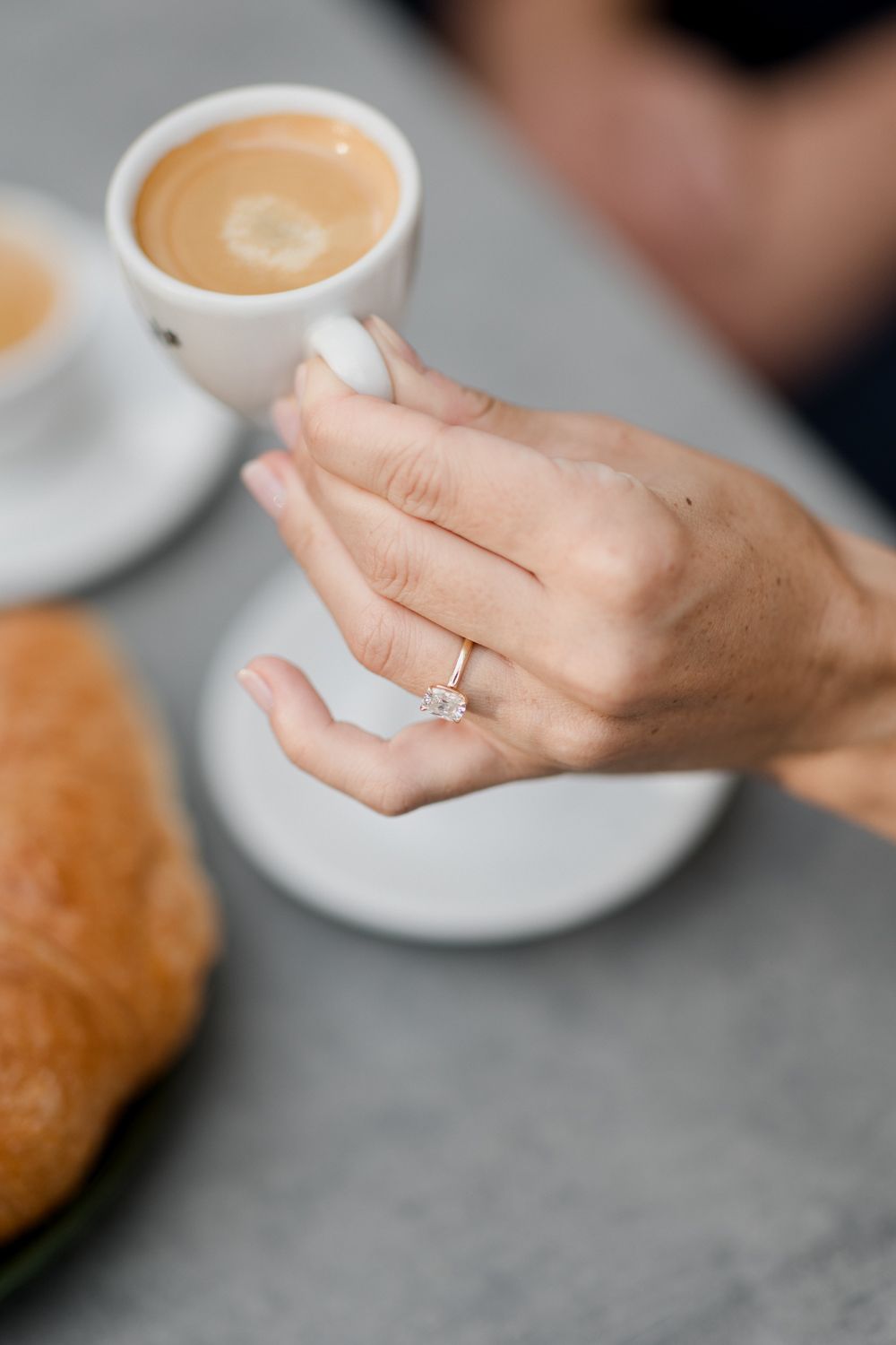Hands hold a delicate white coffee cup with creamy latte art on a grey stone surface next to a fresh baked pastry. Seance couple Tours
