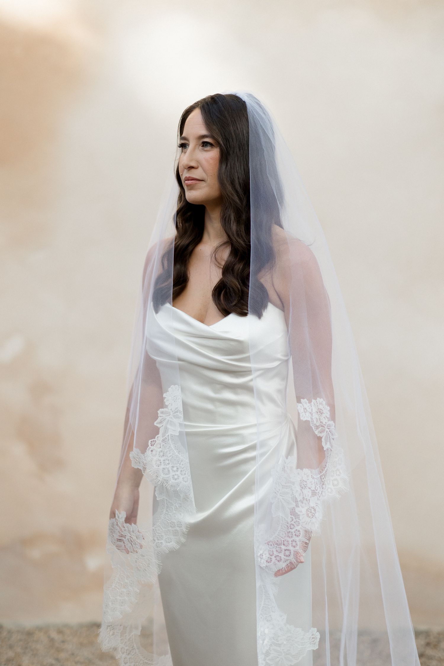 A bride in an elegant white satin gown and long veil poses against a light neutral background.