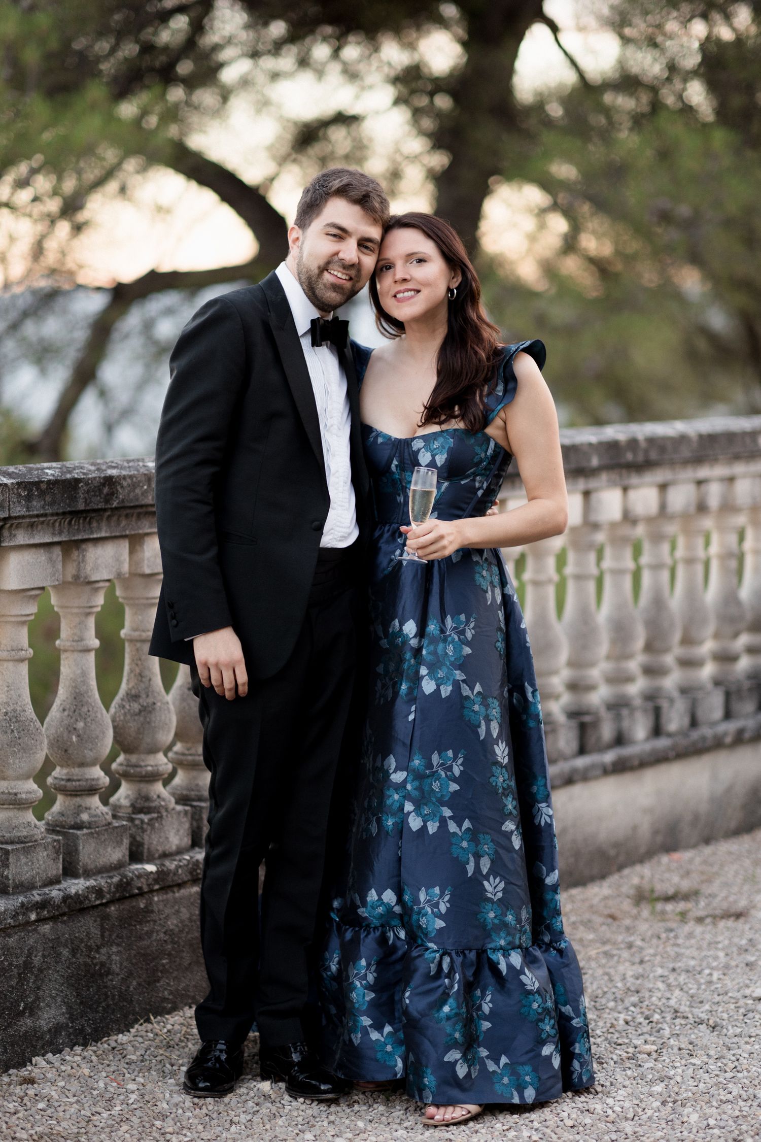A couple in formal attire poses together on a stone balcony with decorative balusters at sunset.