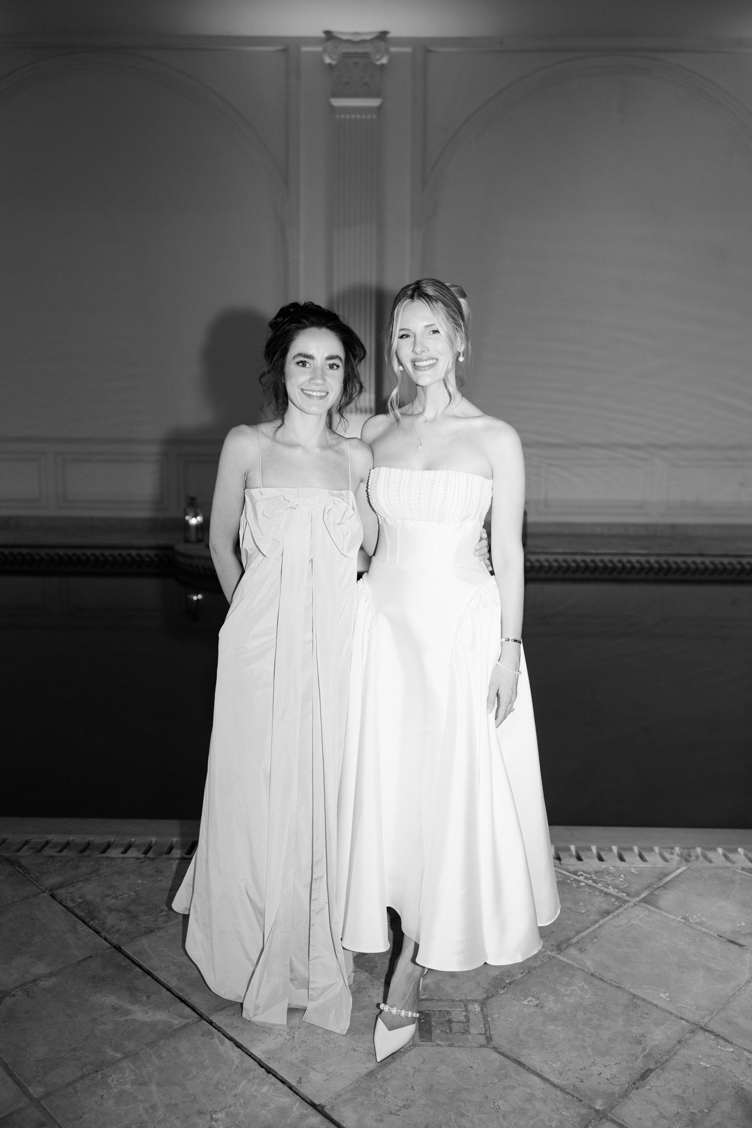 Two figures in white dresses stand together in a black and white photograph series on a tiled floor.