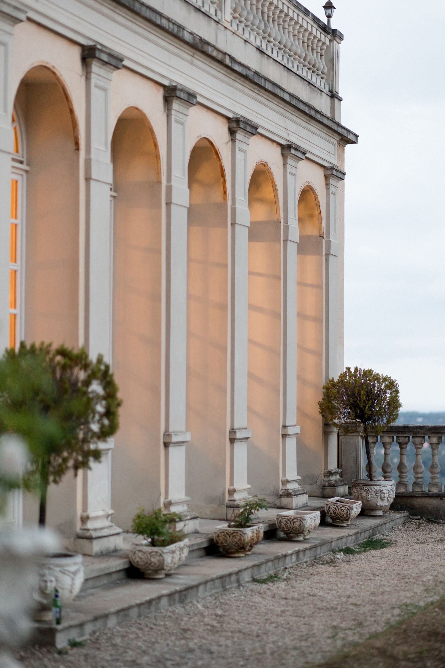 Classical stone colonnade with arched walkway and decorative planters overlooking a scenic view at dusk.
