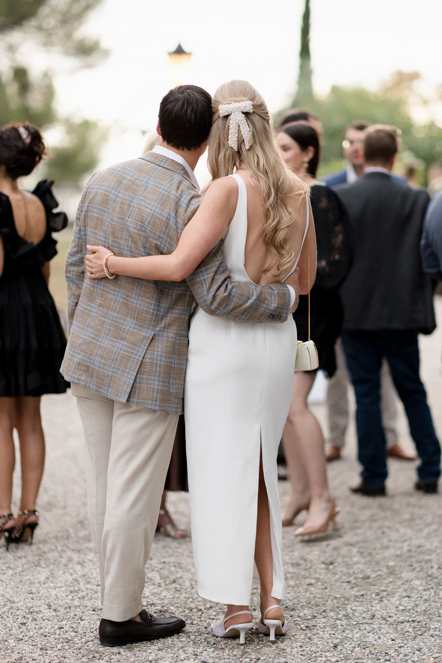 A couple in formal attire embrace while standing among wedding guests on a gravel path at dusk.