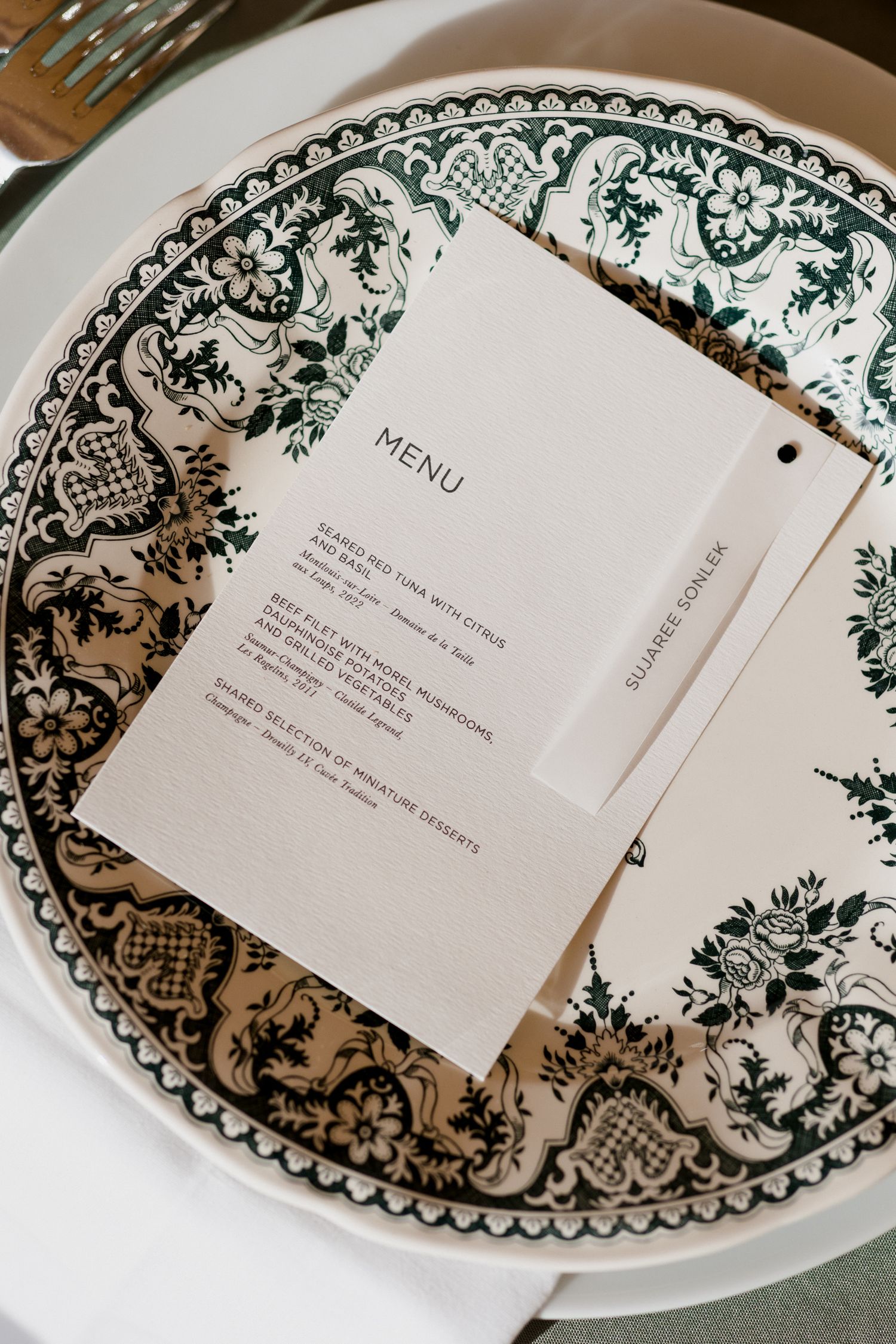 White dinner plates with ornate green floral border designs and menu cards displayed in an elegant table setting arrangement.