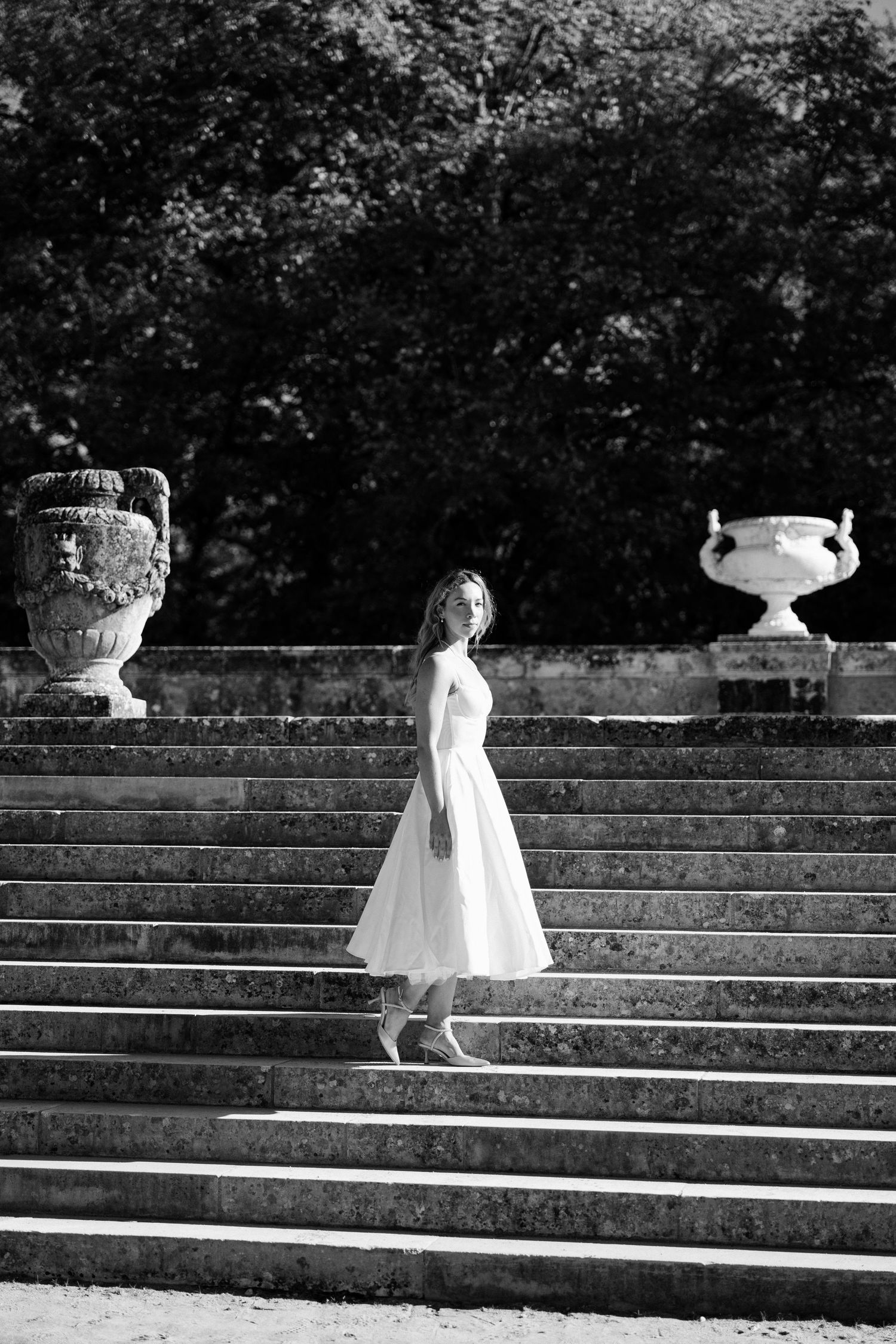 Bride in white tea-length dress stands on grand stone staircase flanked by ornate urns.