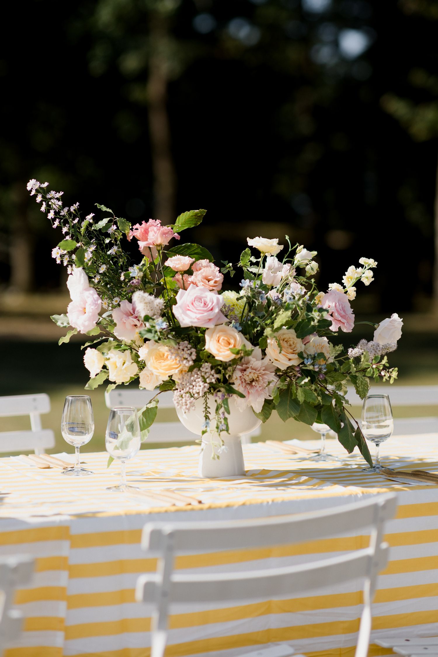 White and pink floral centerpieces in small vases line a table with yellow and white striped tablecloth at an outdoor event.