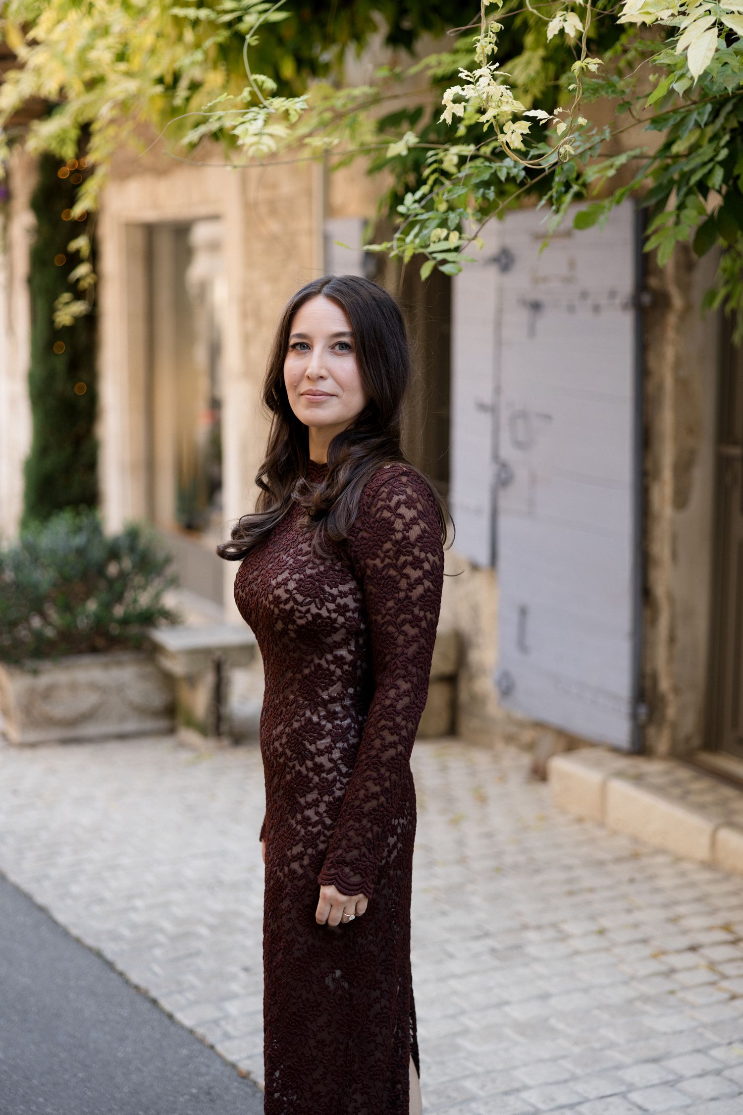 A person wearing a burgundy lace dress poses in a charming cobblestone alley with stone walls and greenery. Engagement session France