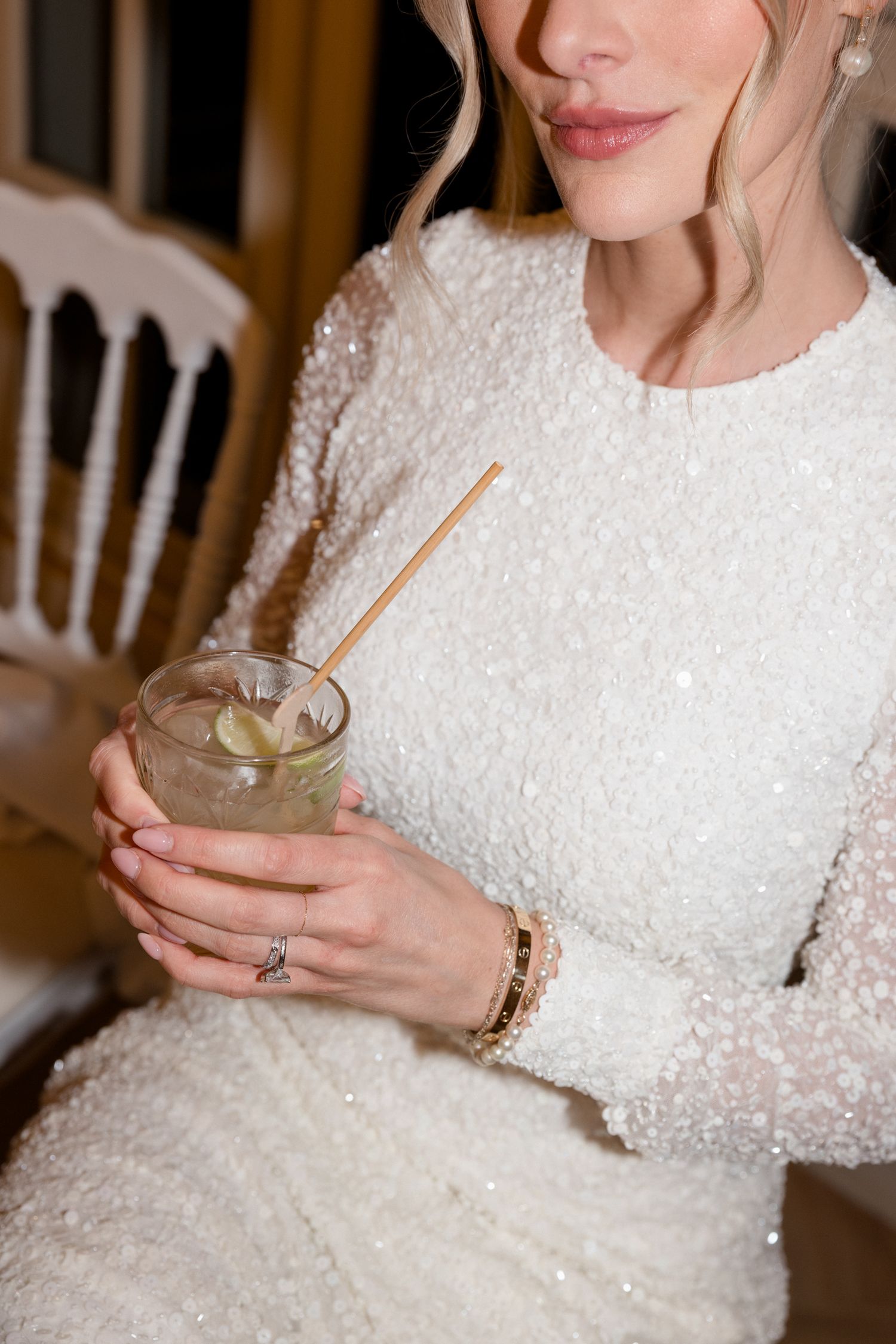 Person in a sparkly white sequined dress holds a glass drink with a gold straw in elegant indoor setting.