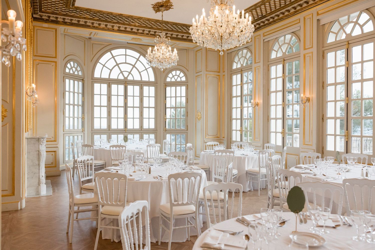 Elegant white ballroom with crystal chandeliers, arched windows, and round dining tables set for a formal event. mariage grasse