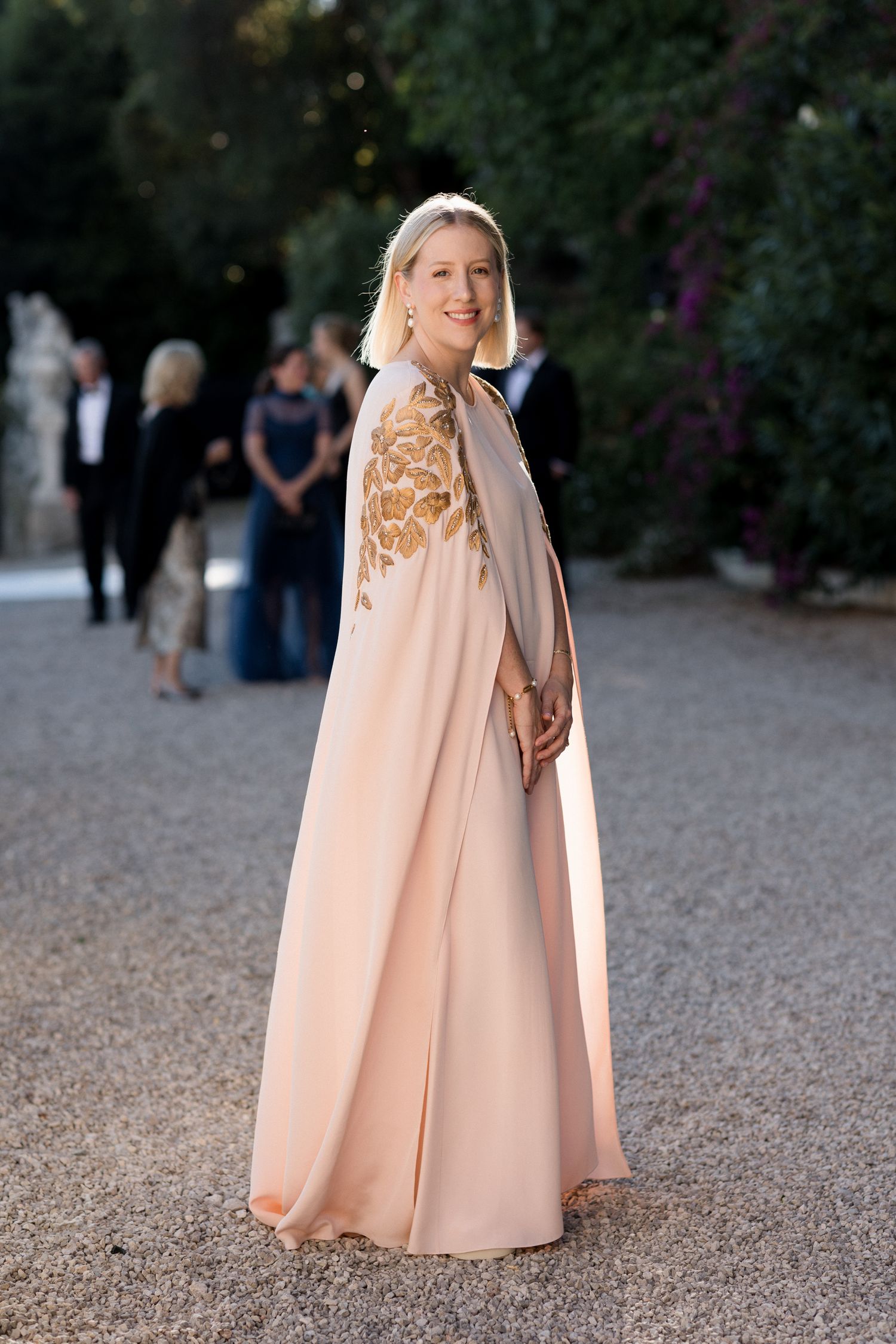 A figure in a flowing peach-colored cape stands on a gravel path at dusk.