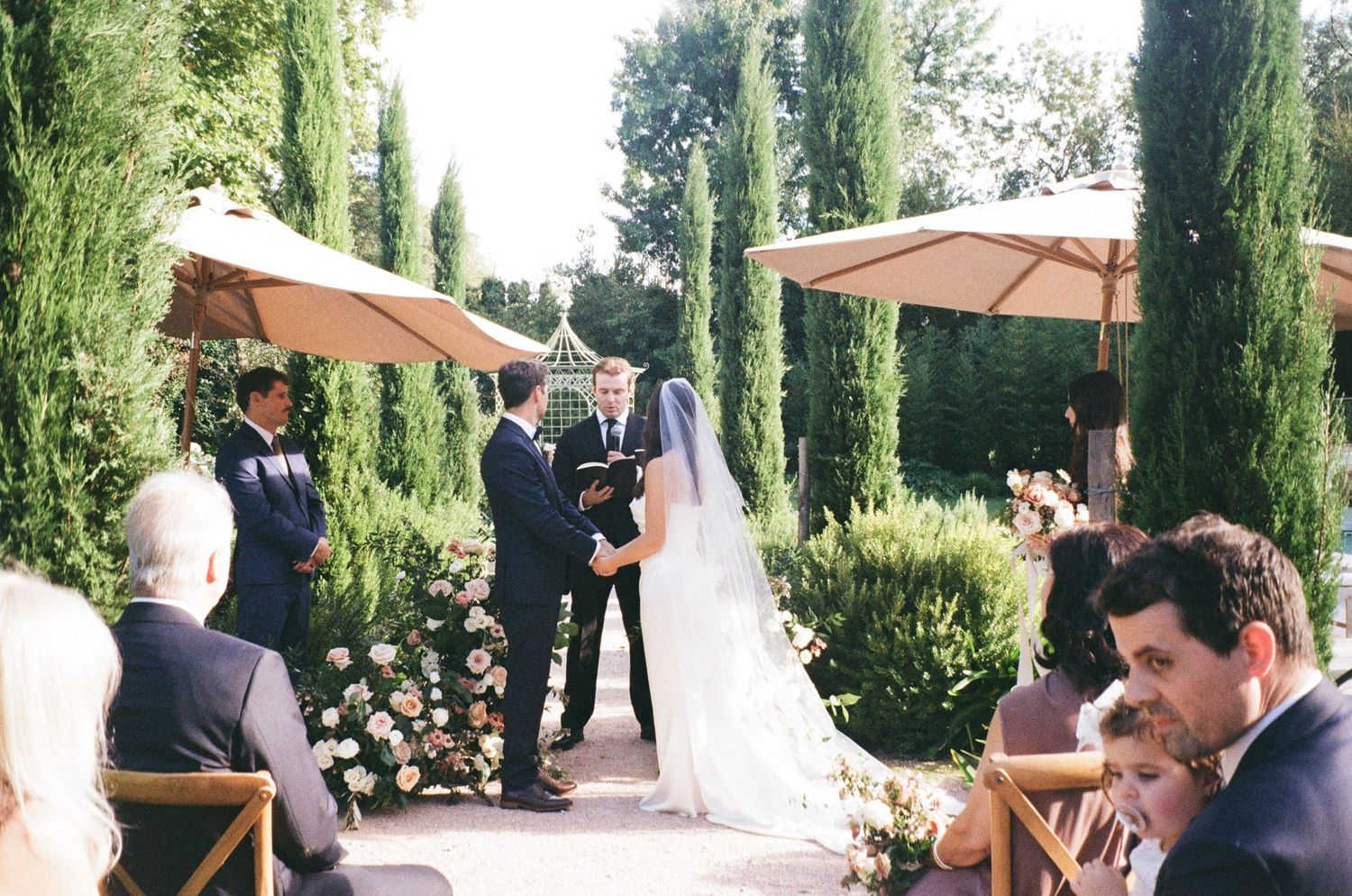Wedding guests mingle and celebrate in an outdoor garden setting with tall cypress trees and patio umbrellas.