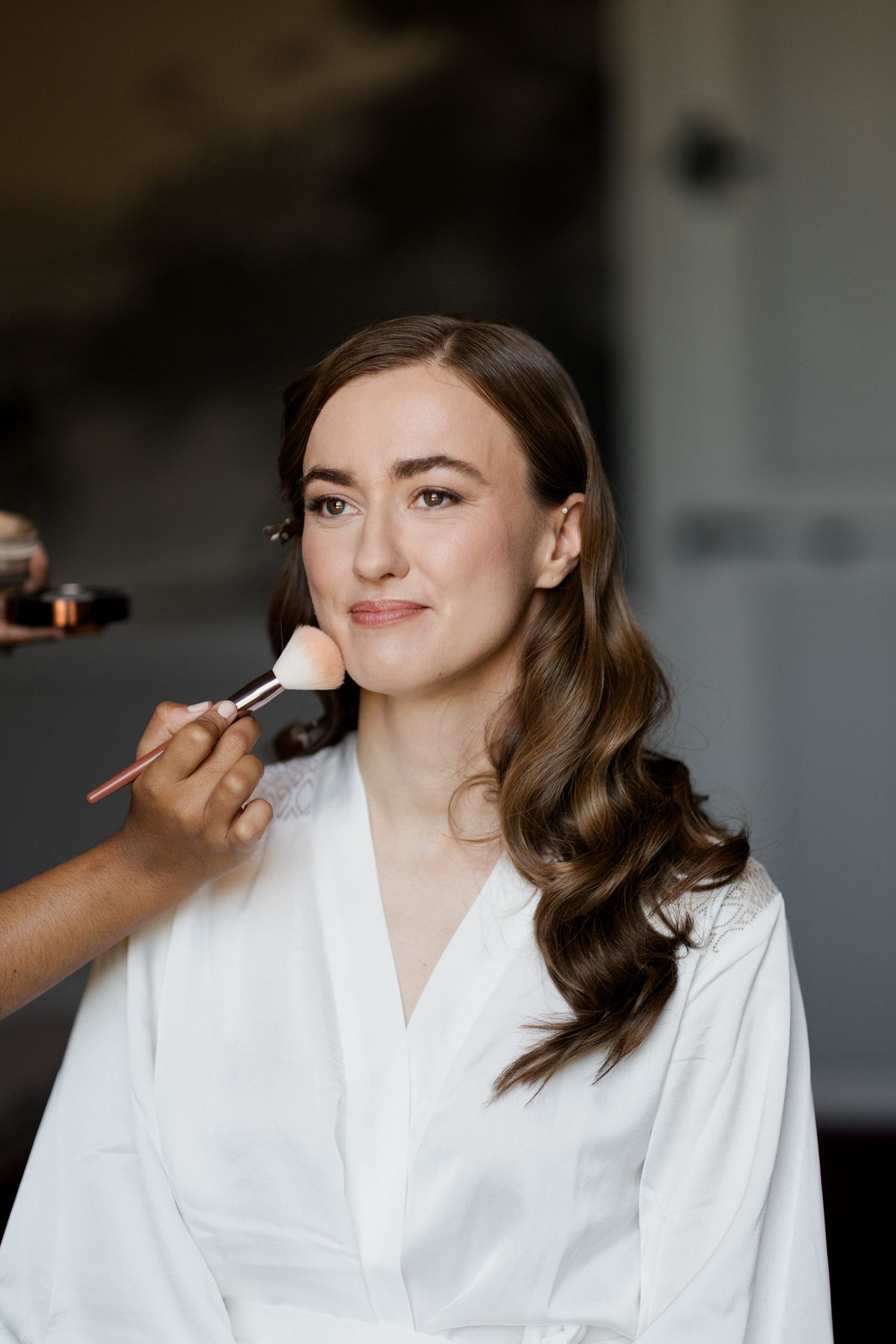 A woman in a white robe gets her makeup done while sitting in a bridal preparation setting. Paris wedding photographer