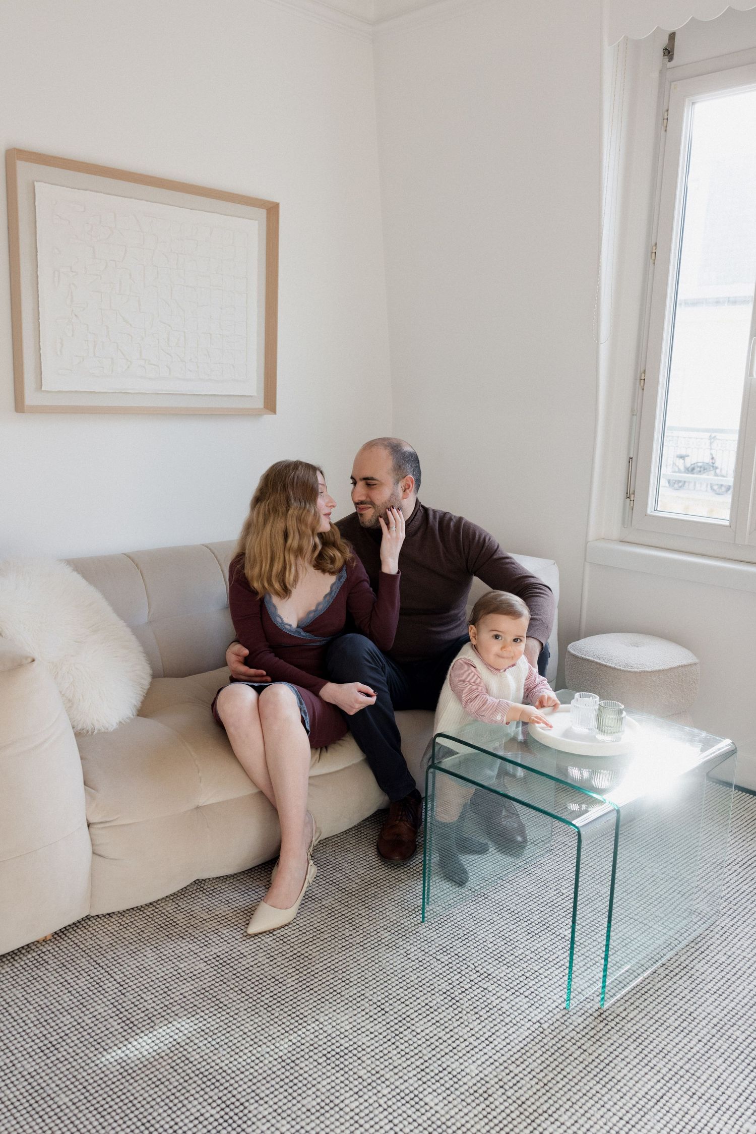 A couple sits closely on a beige sofa in a bright living room while a toddler stands by a glass coffee table.