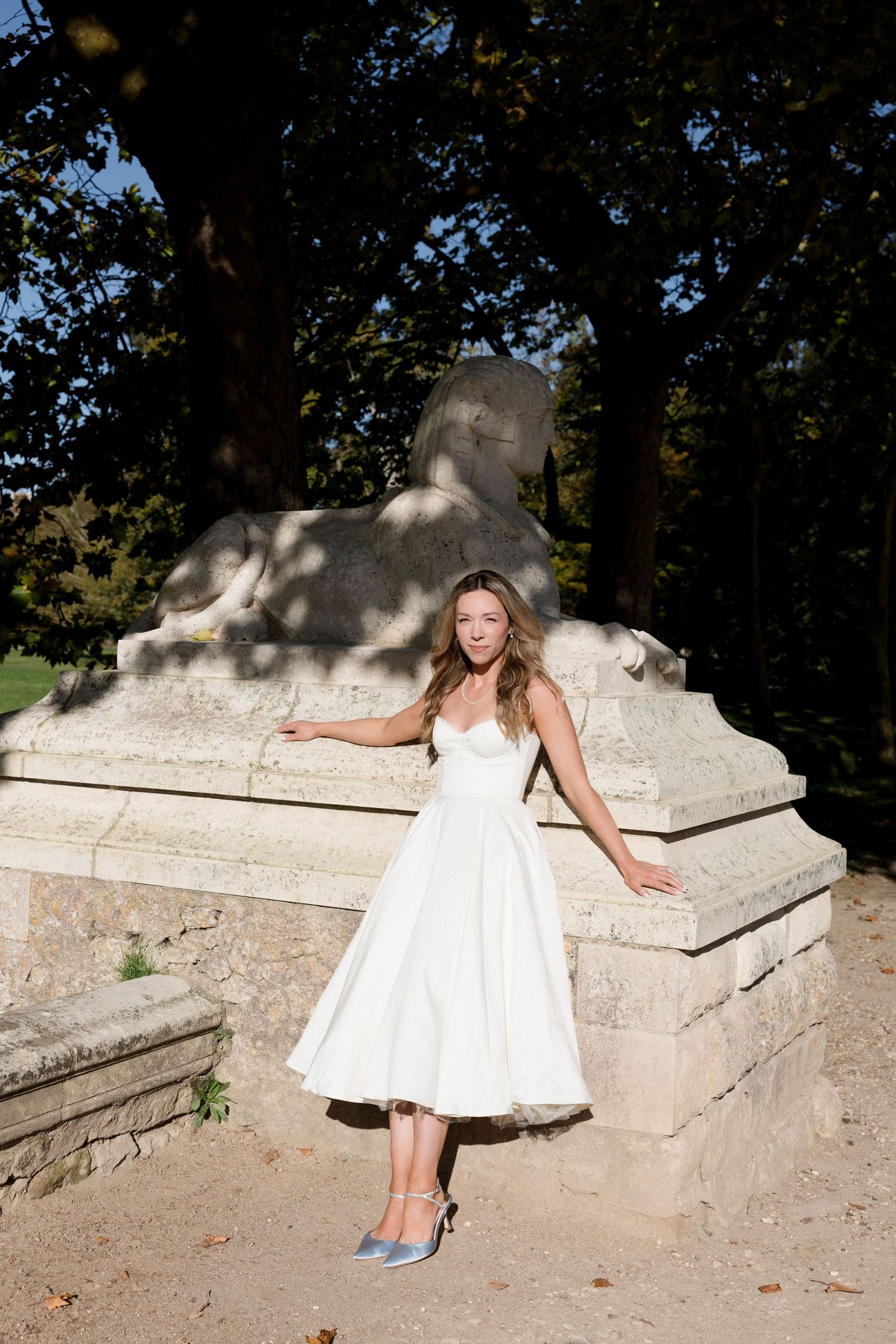Elegant bride in white tea-length wedding dress posing by stone lion sculpture in sunny garden.