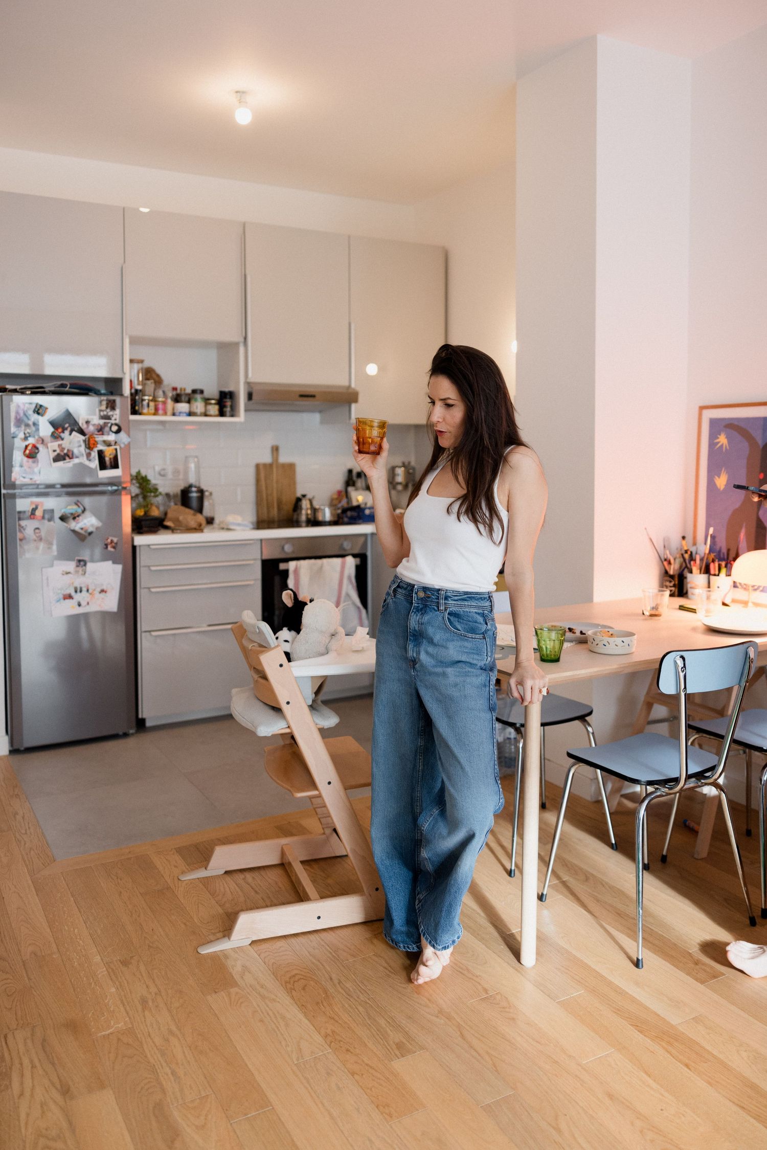 Woman in wide-leg jeans and white tank top stands in modern apartment kitchen with hardwood floors.