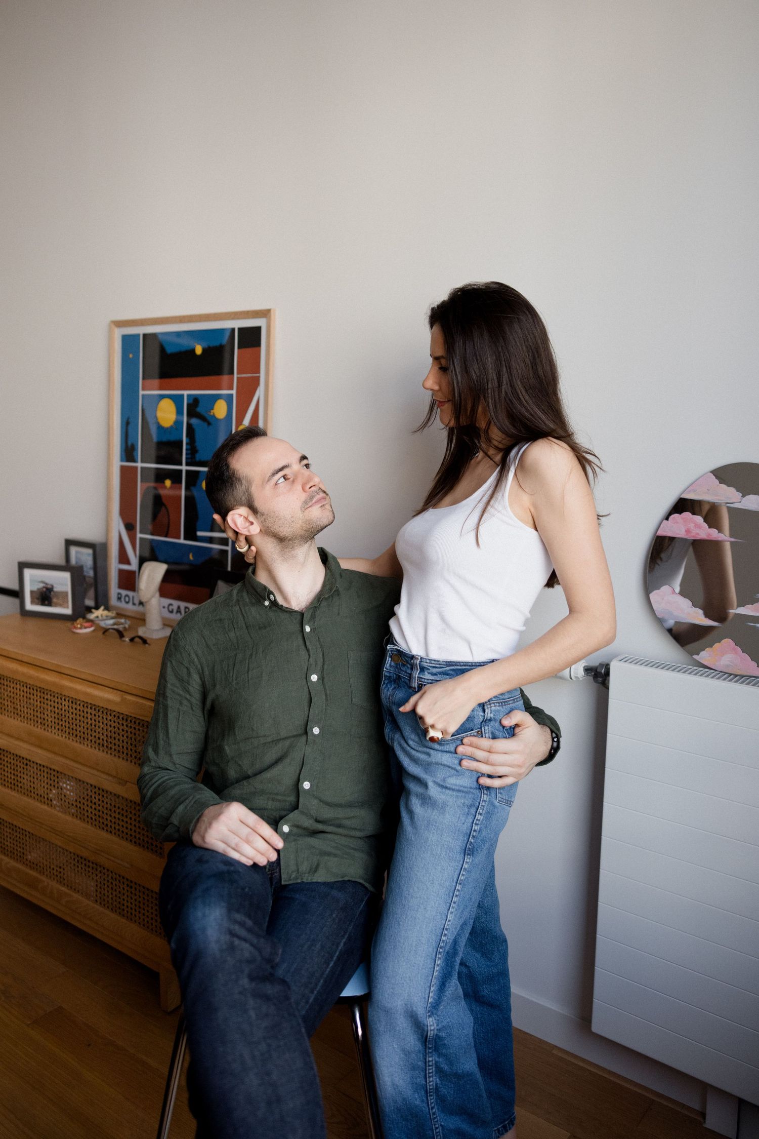 Young couple sharing a tender moment at home, man seated looking up at woman standing beside wooden dresser.