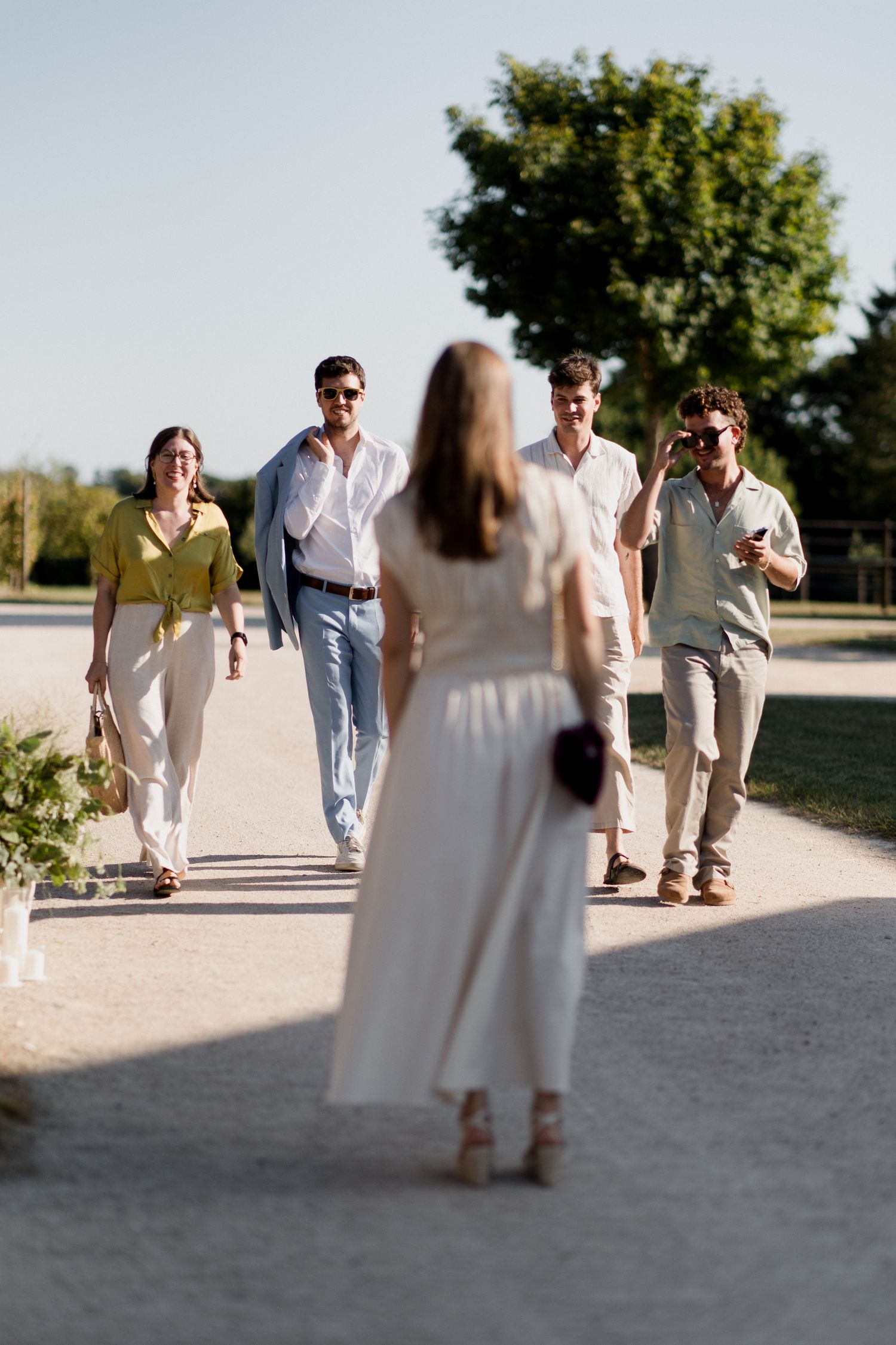 A group of people in light colored casual clothing walk together on a dirt path during a sunny day.