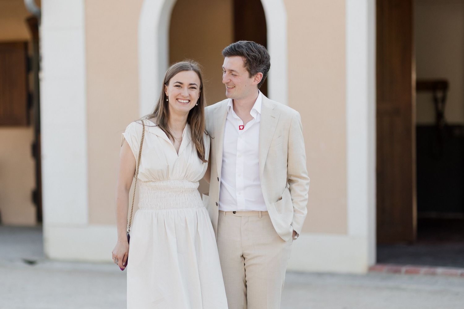 Couple in white and beige outfits walking together in an elegant outdoor colonnade setting.