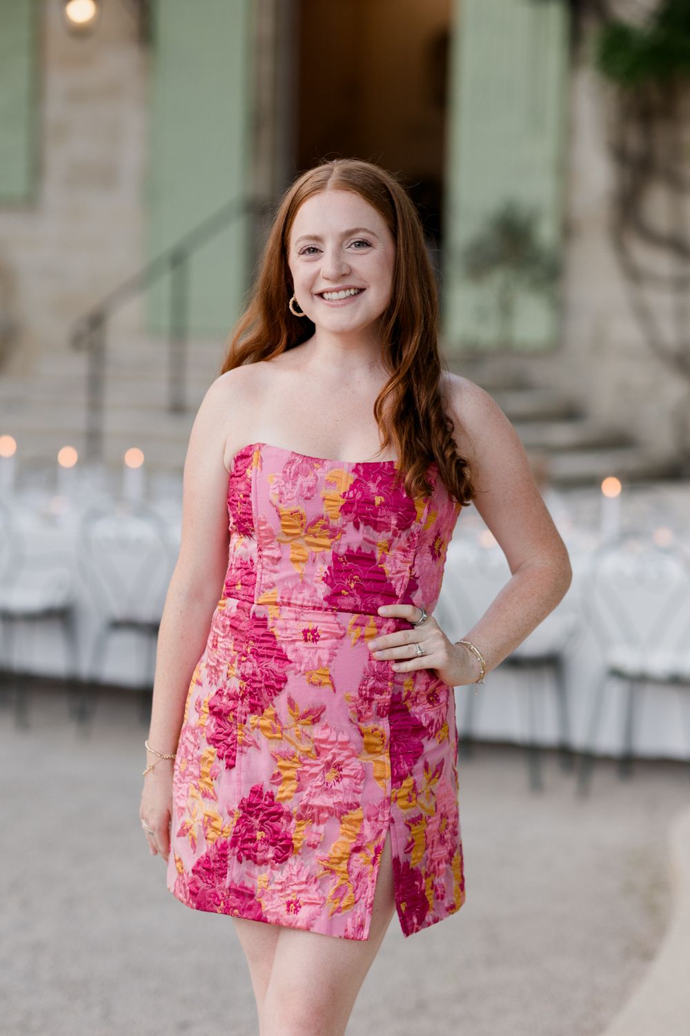 A woman in a floral pink strapless mini dress poses outdoors at an event venue with chairs in the background.