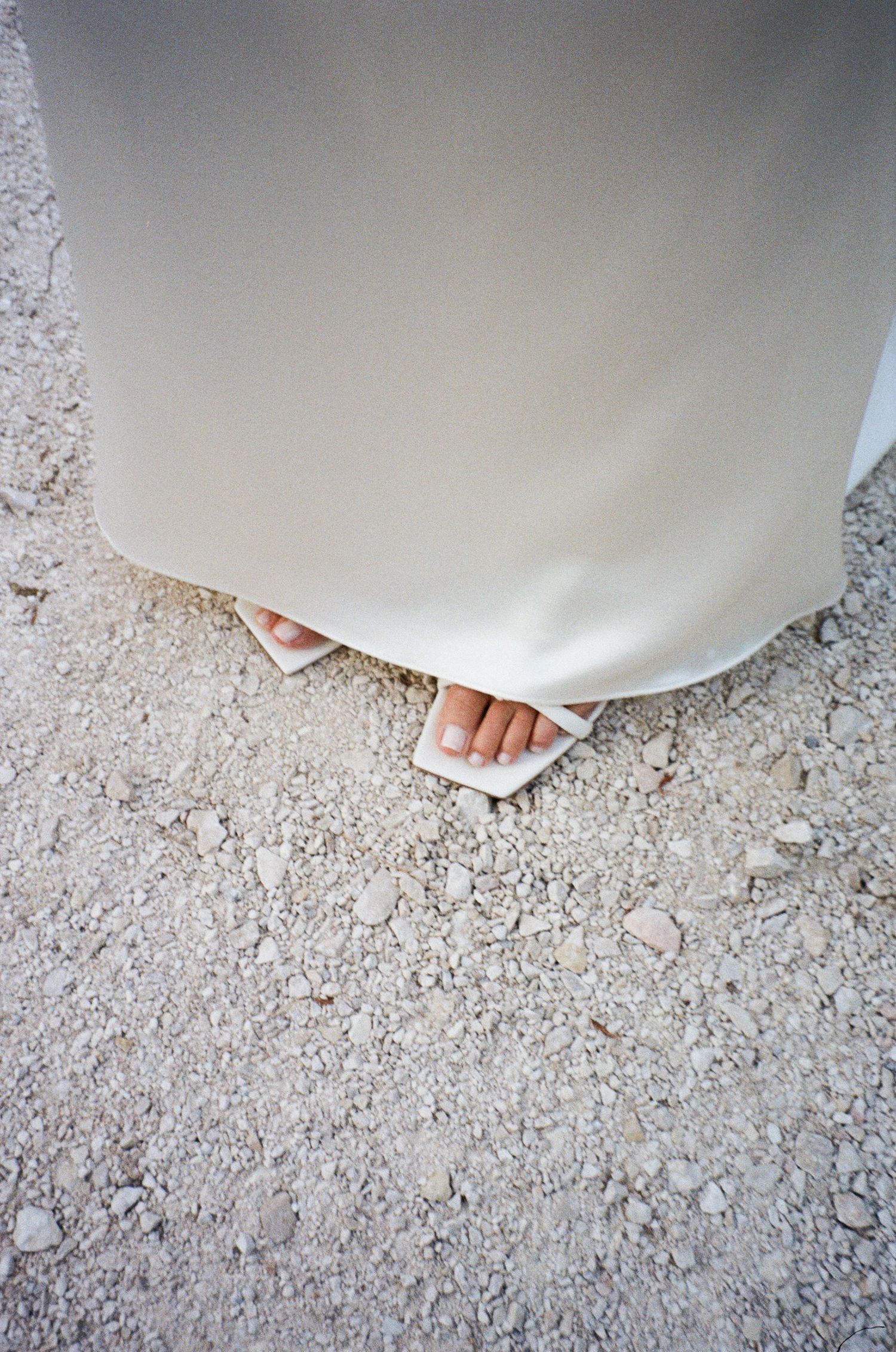 The hem of a white dress or gown brushes against a rough textured ground surface.