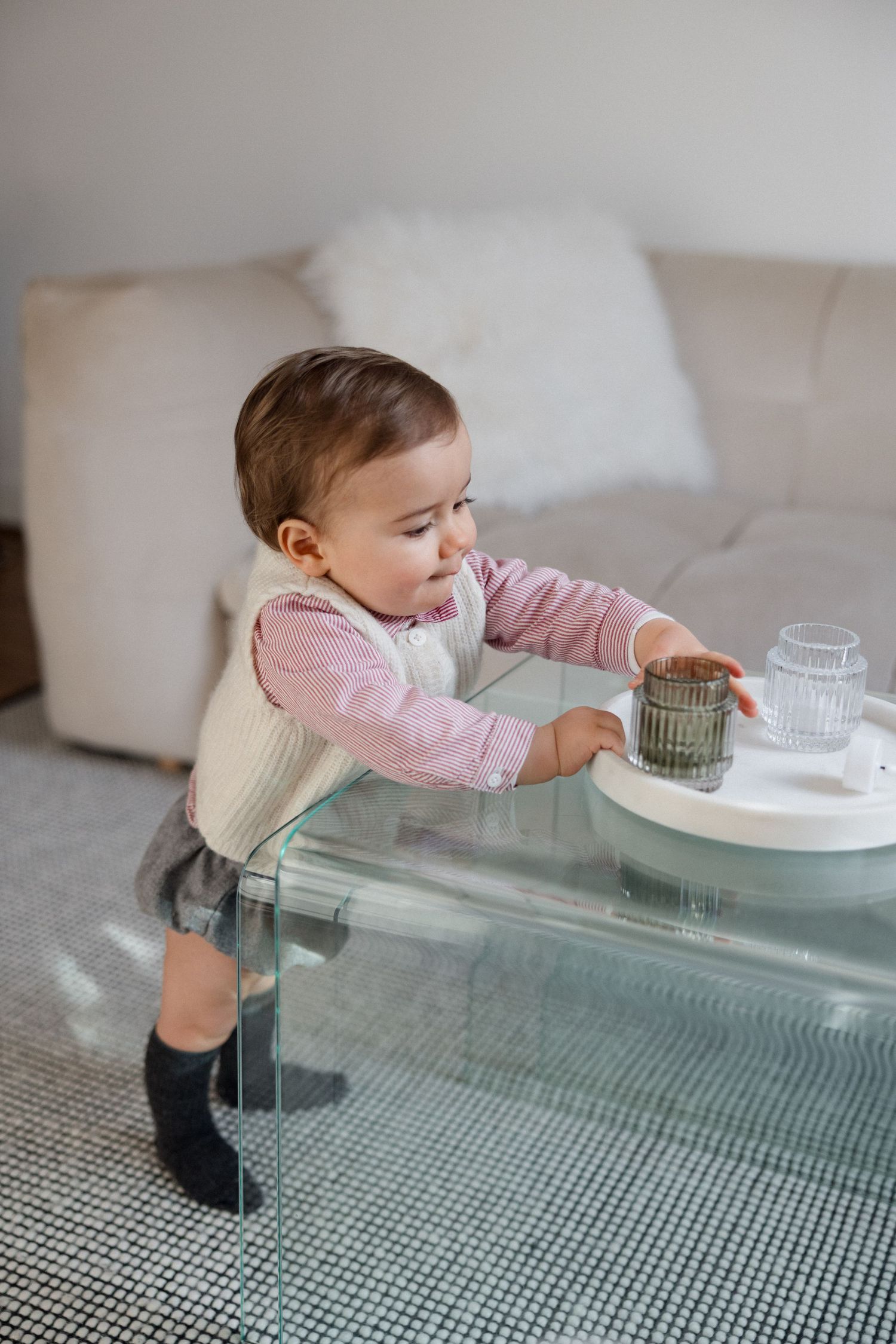 Curious baby in pink shirt reaches for glasses on a glass coffee table, standing beside a white sofa.