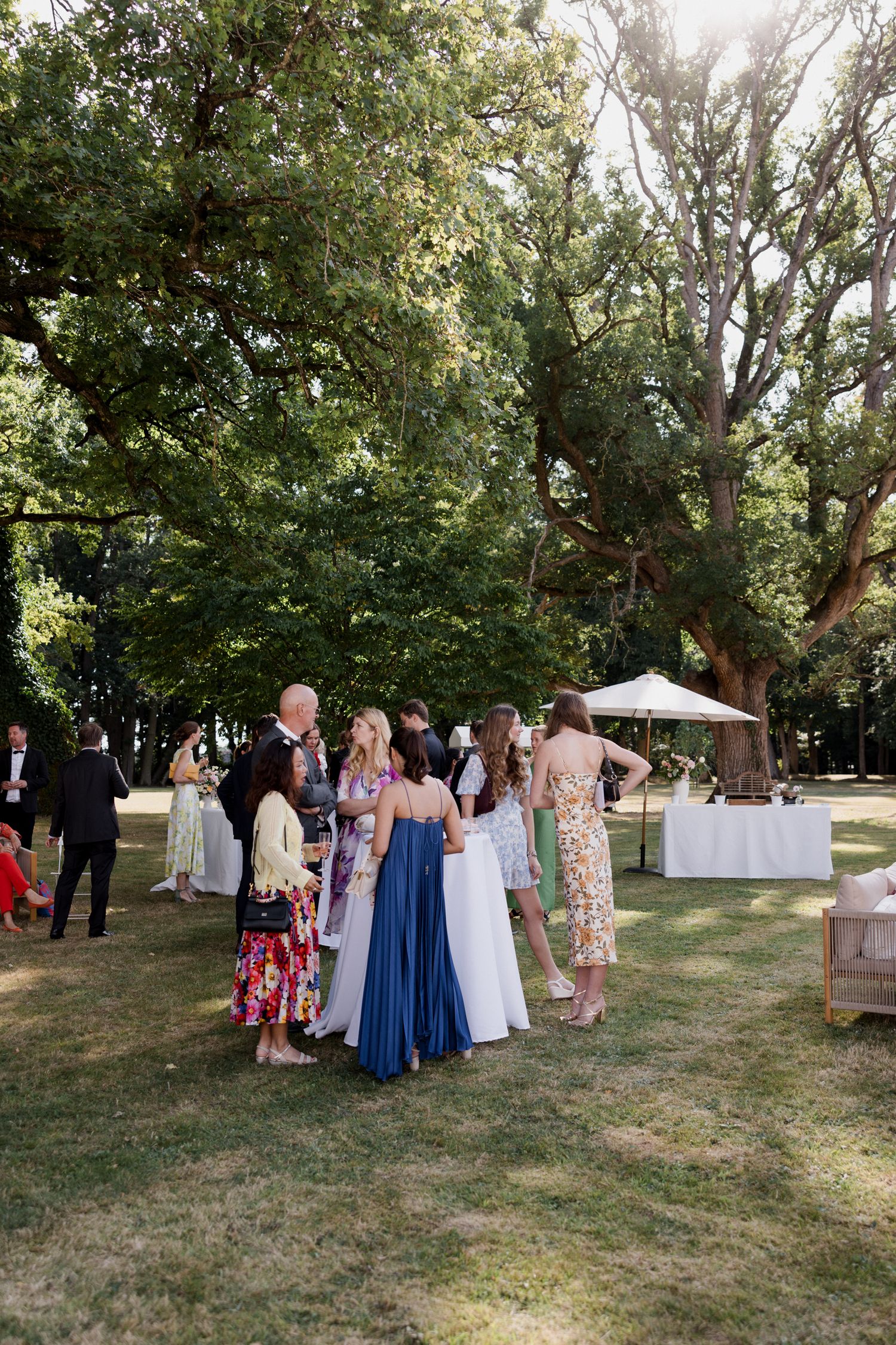 Guests gather outdoors at a garden wedding reception with white tables and umbrellas under tall trees. Paris wedding photographer