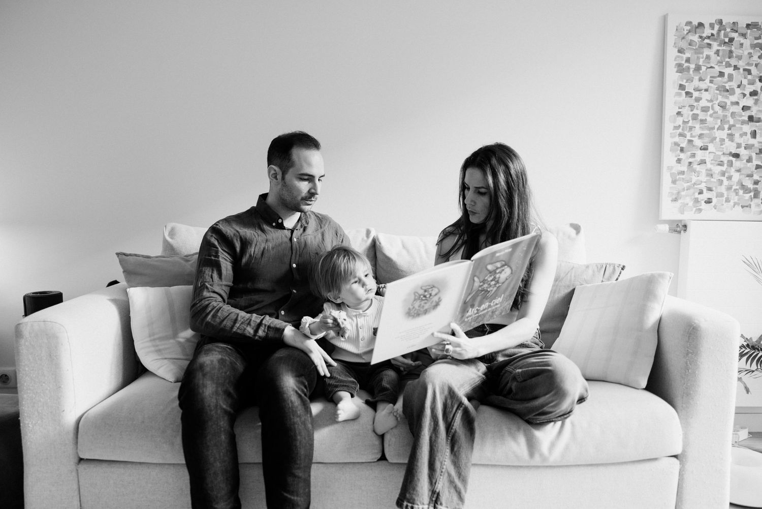 Family of three sitting on a couch reading a picture book together in a cozy living room, black and white photo.