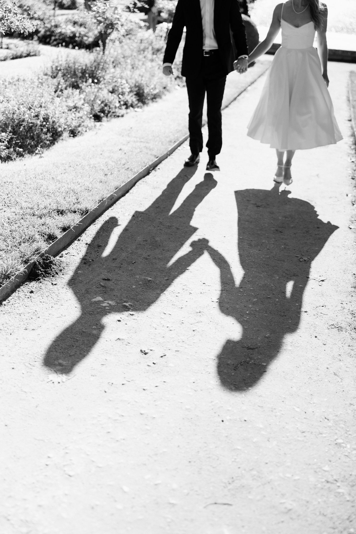 Black and white photo of newlyweds holding hands walking on a path with their shadows cast below.