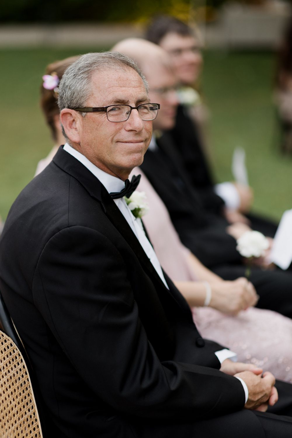 Person in a black tuxedo and glasses seated at an outdoor event during what appears to be a wedding ceremony.