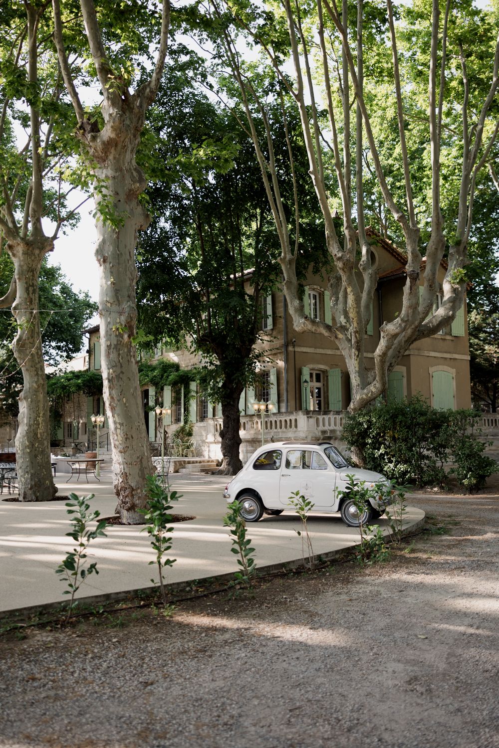 Vintage white Fiat 500 parked on tree-lined street beside historic Mediterranean villa in dappled sunlight.