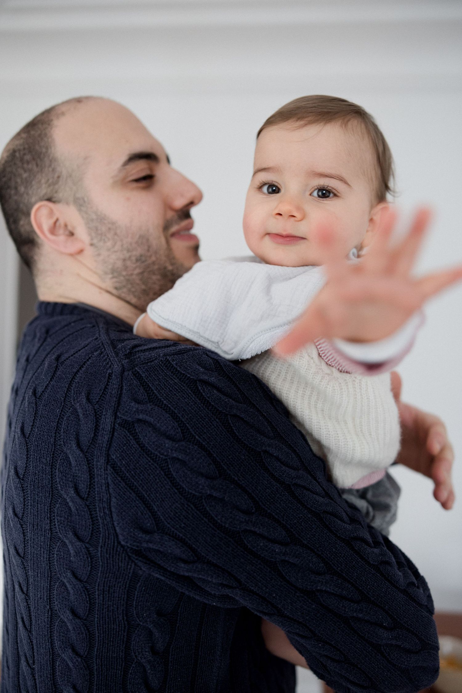 A bearded man in a navy cable-knit sweater holds a curious baby reaching toward the camera in a bright white room.