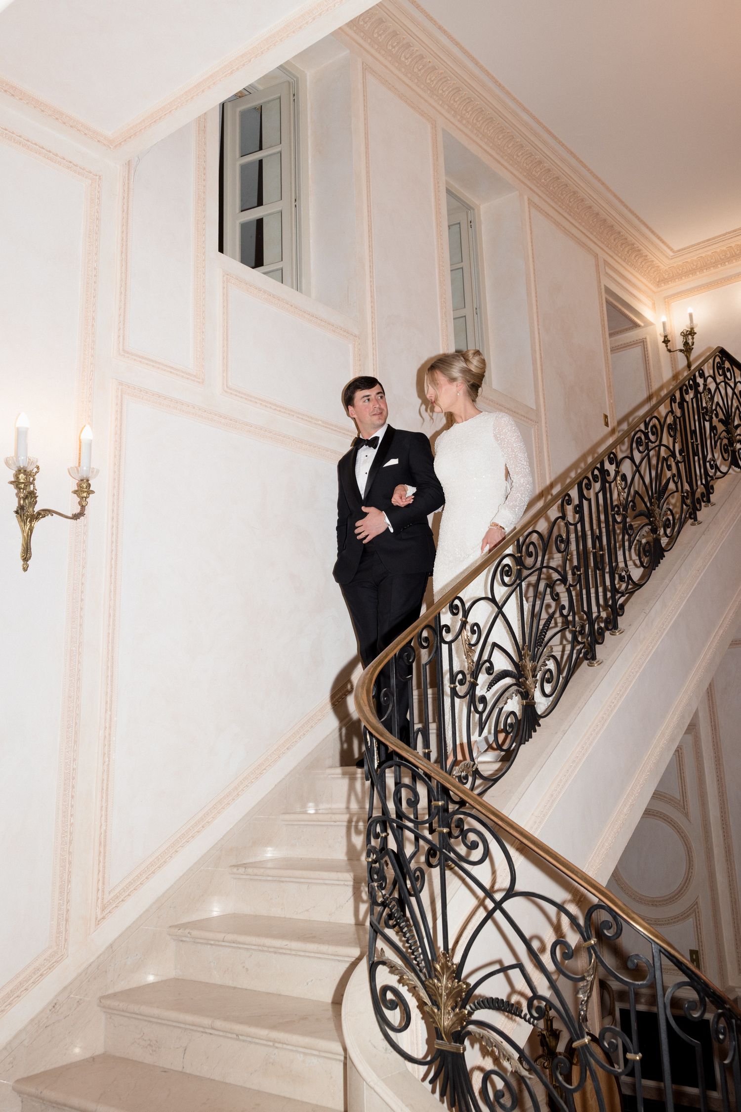 A couple descends an elegant staircase with ornate wrought iron railings in a grand white interior setting.
