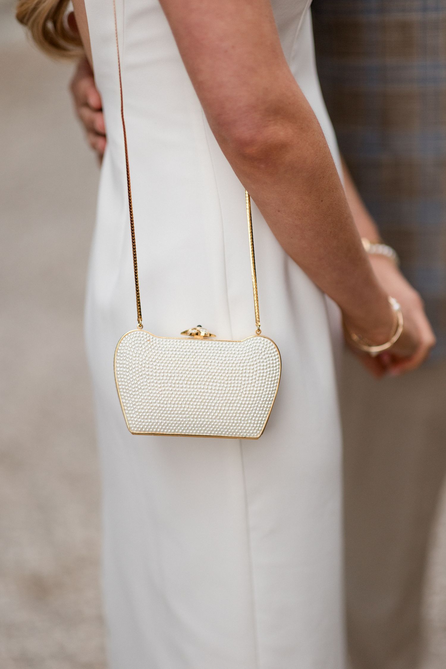 A small white textured clutch purse with gold chain strap hangs elegantly against a flowing white dress. destination wedding france