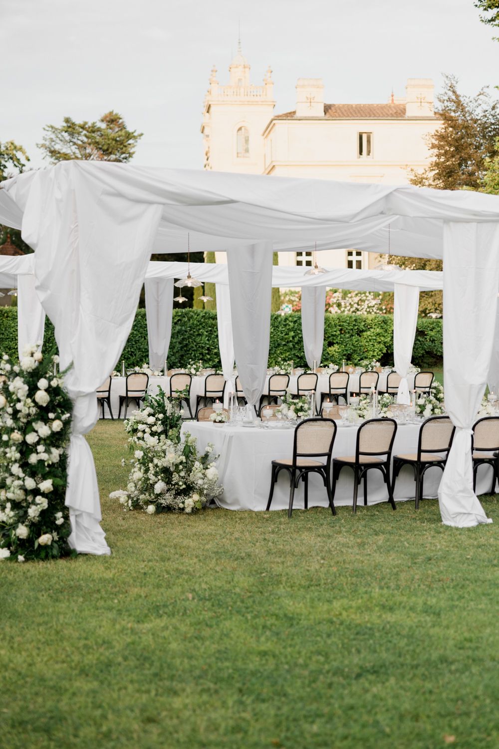 Elegant outdoor wedding ceremony setup with flowing white drapes, dark chairs, and white floral arrangements on the lawn.