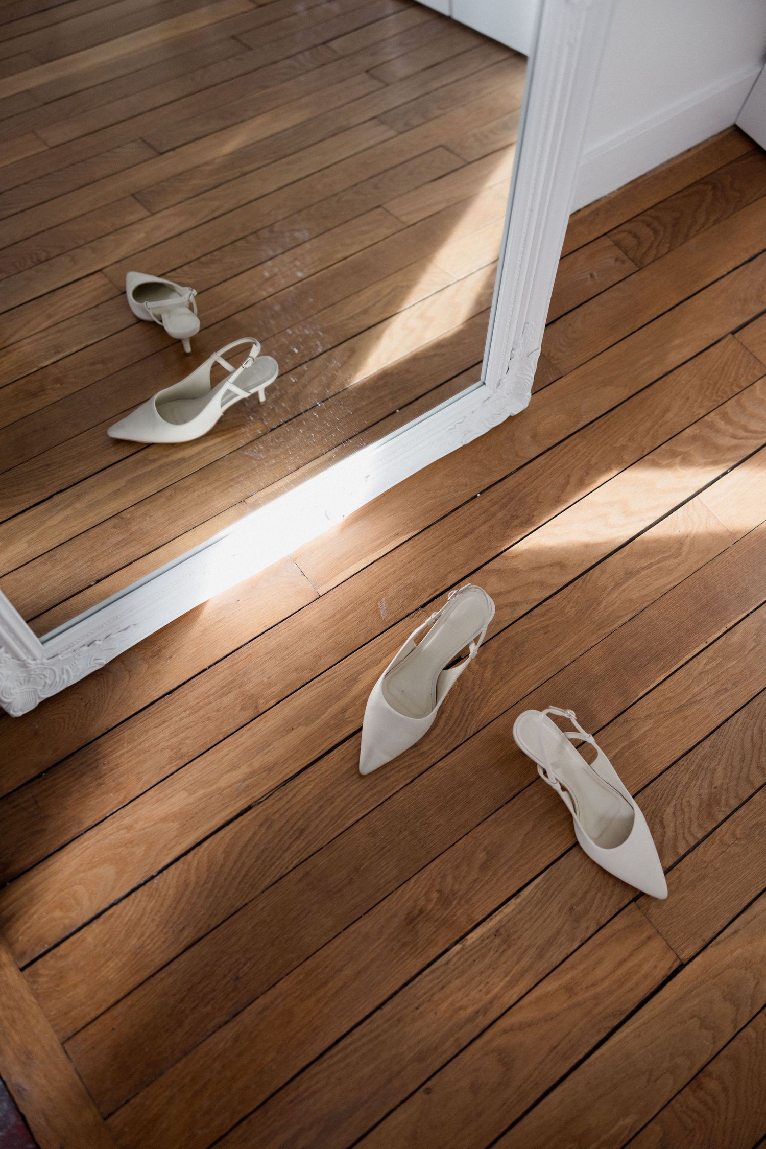 White slingback heels on a wooden floor reflected in a large mirror in a sunlit room.