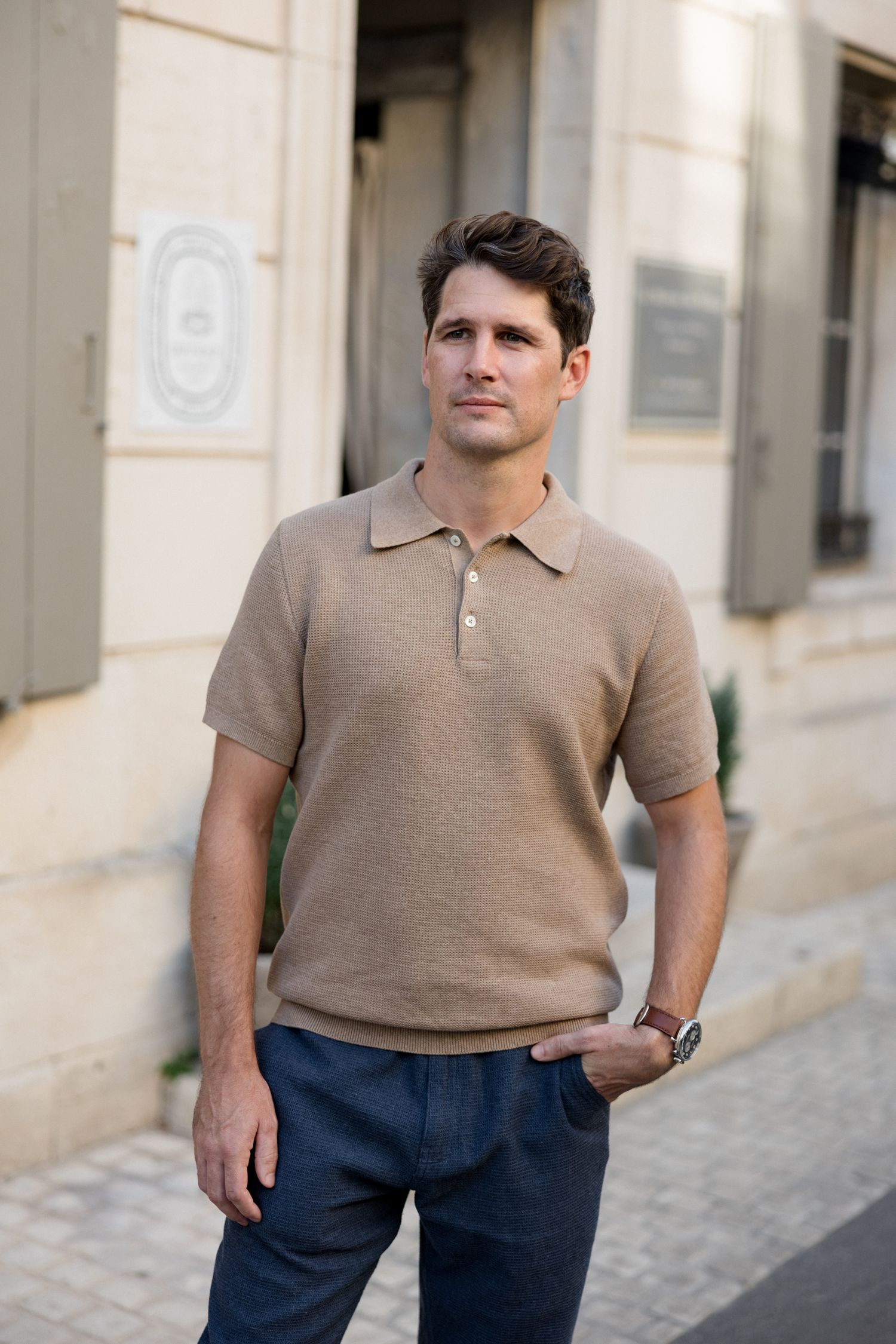 Person wearing a beige knit polo shirt and navy pants poses casually on a cobblestone street.